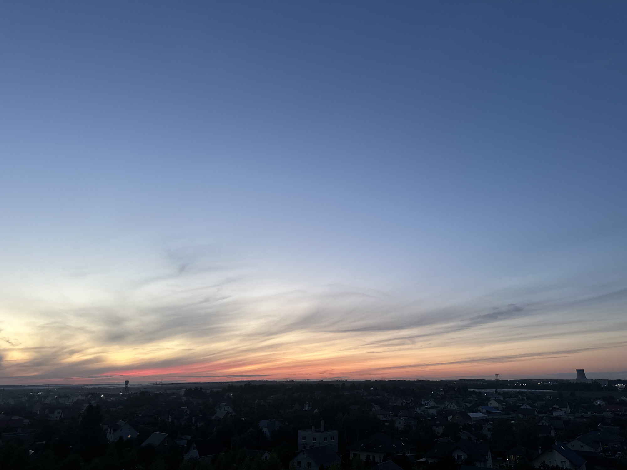 The image captures a panoramic view of a residential area at dusk. The sky transitions from a deep blue at the top to a gradient of lighter blues, pinks, and oranges near the horizon, indicating the setting or rising sun. Wispy clouds are scattered across the sky, illuminated by the fading light. Below the sky, a dense landscape of houses with dark roofs stretches across the frame. The houses appear to be mostly two-story structures with visible windows and some with illuminated interiors. Trees and vegetation are interspersed throughout the residential area, contributing to the slightly silhouetted effect of the foreground and midground. In the distance, some taller structures, possibly industrial or communication towers, are visible, along with what appears to be a cooling tower on the far right. The overall scene is one of a quiet, suburban or rural community settling in for the evening under a colorful sky. The location is identified as Shchomyslitski Sel'ski Sovet, Belarus. There are no visible people or ongoing actions depicted in the image.