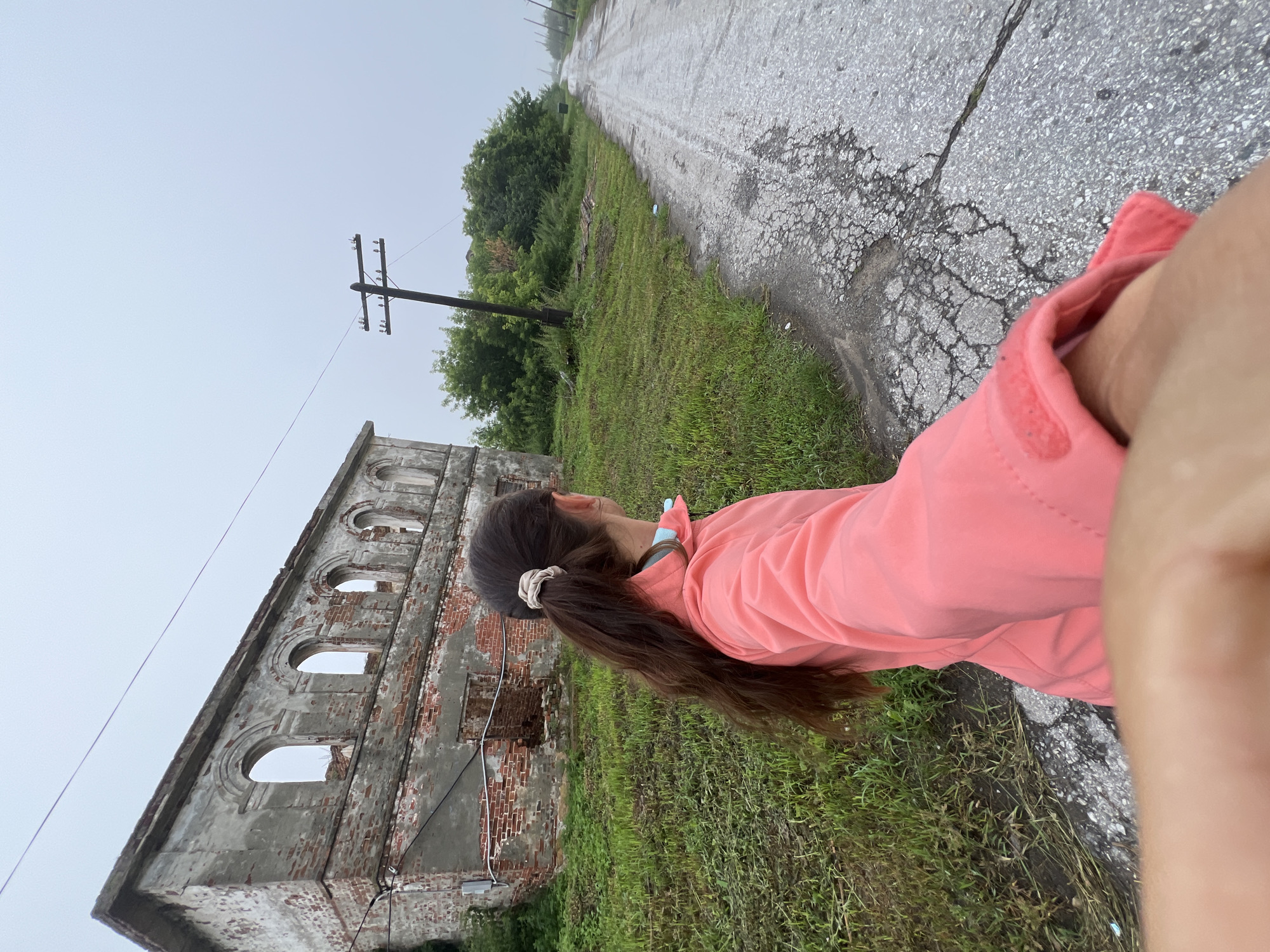 The image depicts a person from behind, walking along a cracked asphalt road towards an abandoned brick building and a utility pole. The building appears to be in disrepair, with broken windows and exposed brickwork. The surrounding environment is overgrown with grass and trees, suggesting an undeveloped or neglected area. The sky is overcast and hazy, contributing to a somber atmosphere. The overall scene conveys a sense of decay and abandonment in what appears to be a rural or semi-rural setting. It is situated in the city of Pivkino, Russia.