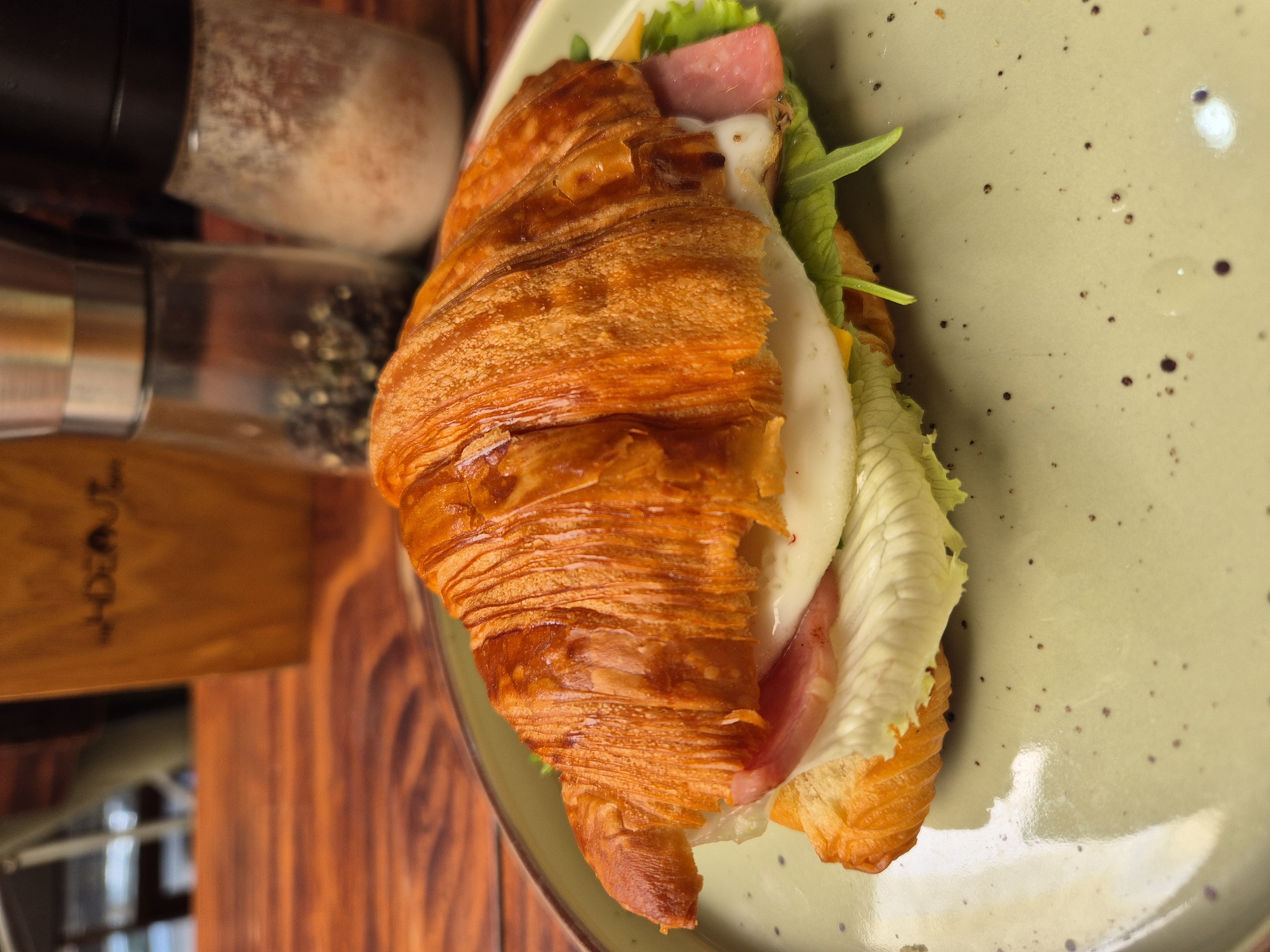 A close-up, eye-level shot shows a croissant sandwich on a speckled, pale green plate. The croissant is golden brown and flaky, split horizontally and filled with a fried egg, ham, cheese, and lettuce. The sandwich is centered on the plate, which is placed on a rustic wooden table. In the blurred background, two salt and pepper grinders and a yellow container with text that appears to read "WHISPER" are visible. The overall scene suggests a casual meal in a cafe or restaurant. The provided context indicates this scene is located in Đà Nẵng, Vietnam.