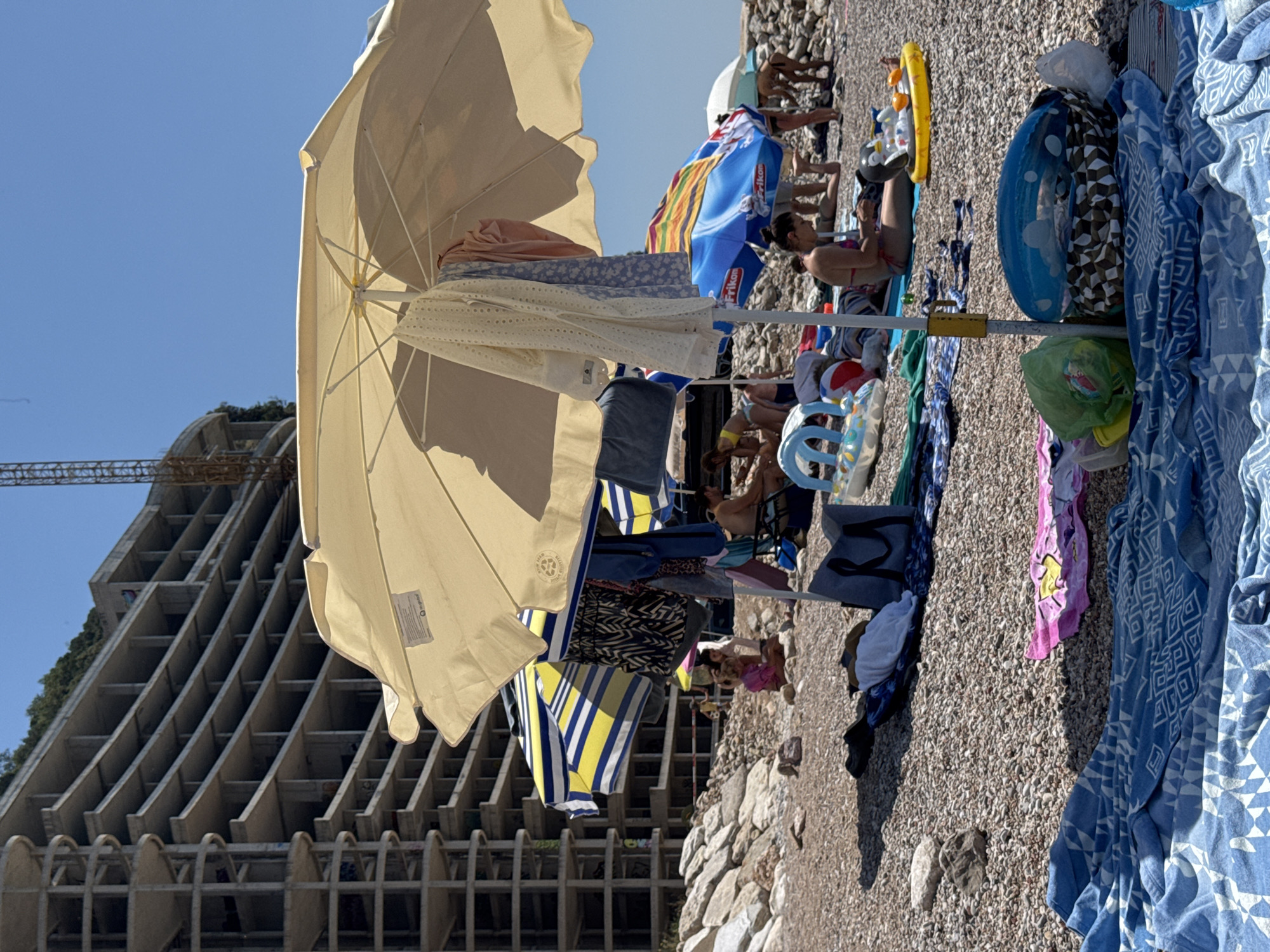 The image depicts a sunny day on a pebbled beach in Perazića Do, Montenegro, with an unfinished, multi-story concrete building under construction in the background. In the foreground, several beach umbrellas are set up, providing shade. One large, cream-colored umbrella is prominently featured, with clothing items hanging from its pole. Beach towels, bags, and inflatable toys are scattered on the ground around the umbrellas. People are visible in the background, some sitting under umbrellas, others potentially swimming or walking. The sea is not directly visible, but the presence of a beach implies its proximity. The overall atmosphere is one of a typical summer beach day, albeit with the unusual backdrop of a large construction project.