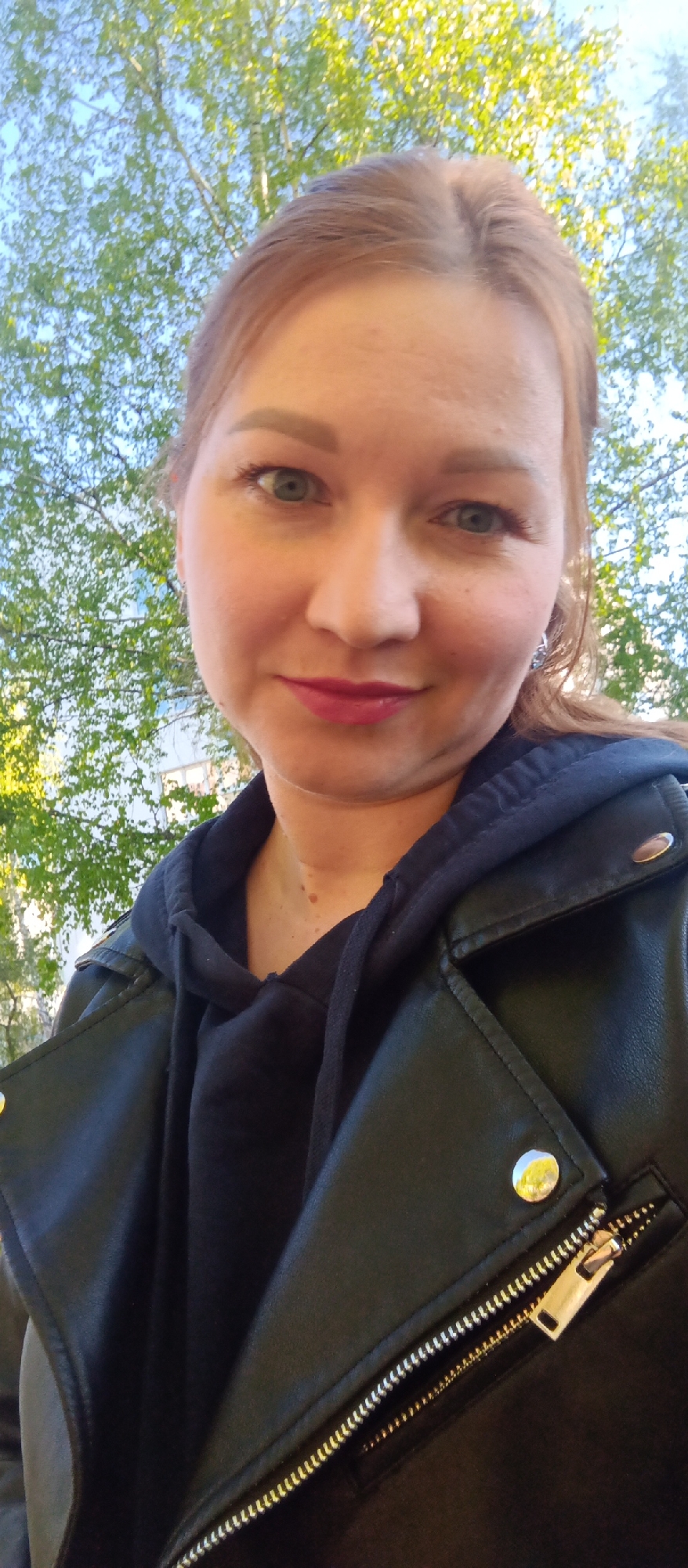 A woman poses for a selfie outside in an unknown city, unknown country. She has fair skin, light brown hair, and green eyes. She is wearing a black hoodie and a black leather jacket. The zipper on the jacket is gold. The background includes green trees and a blue sky with some visible buildings.