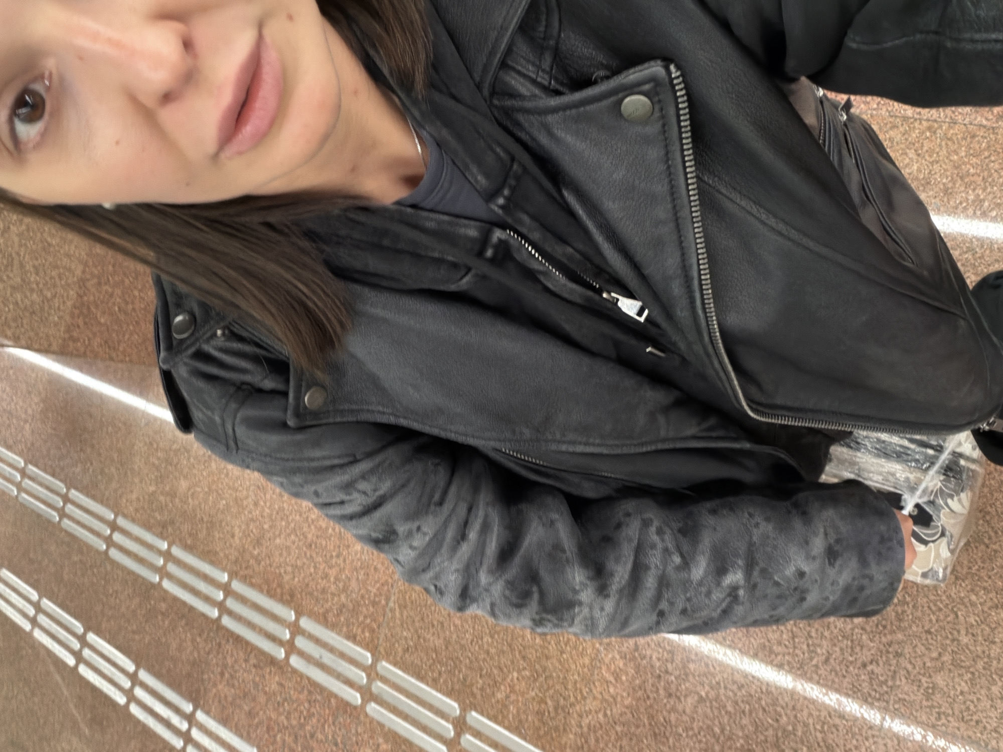 A first-person view shows a person wearing a black leather jacket standing on a brown and white textured surface, possibly indoors. The person's face is visible in the upper left of the frame. The environment appears to be a building interior, possibly a metro station based on the textured surface meant for pedestrians. There is no additional information to determine if this is Moscow, Russia.