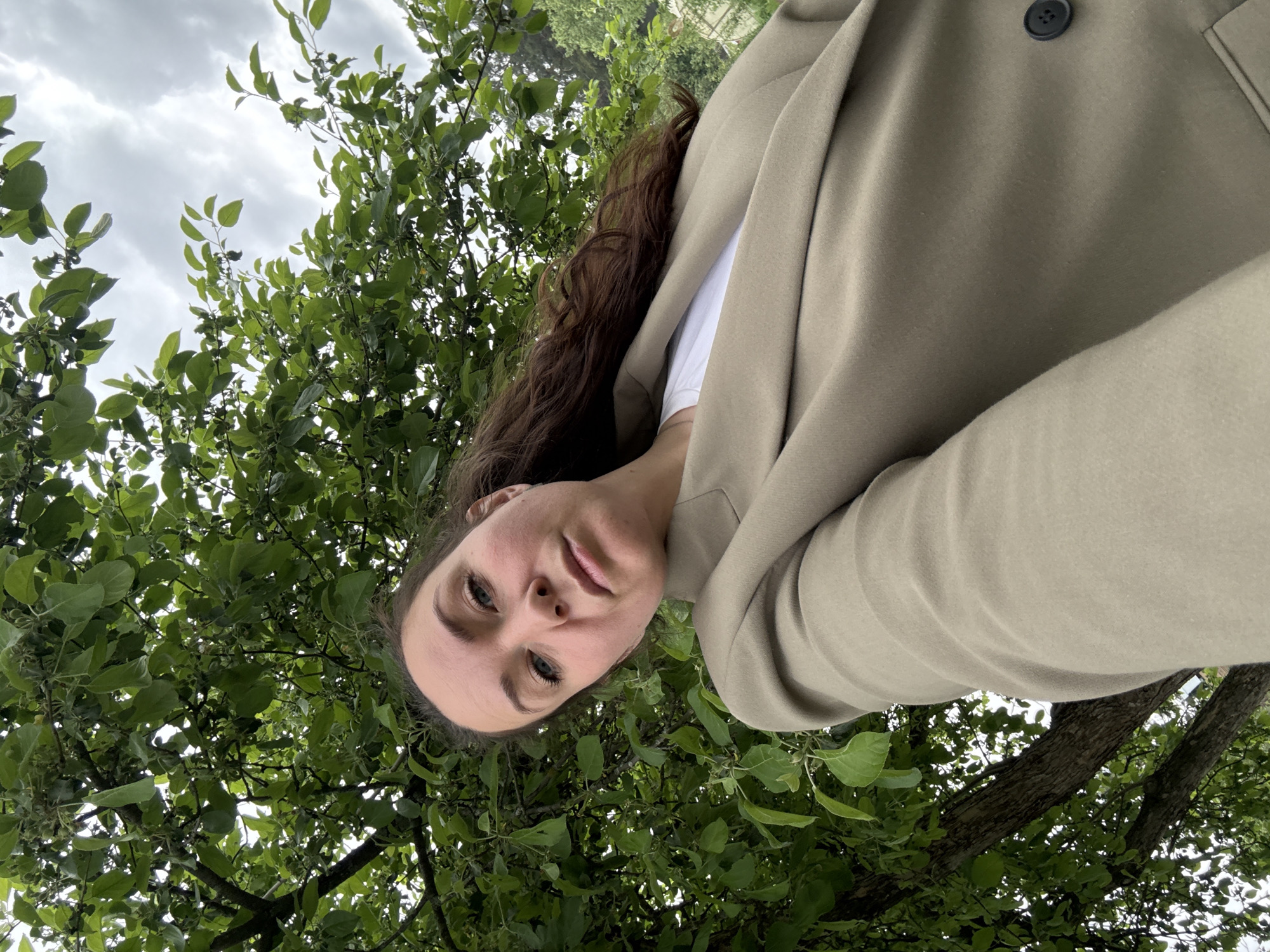 The image shows a woman in a beige blazer and white t-shirt, with long, brown hair. She is outdoors, beneath a tree with green leaves, partially obscuring her face. The sky is visible with cloudy overcast conditions. There is no way to determine if the location is Chaikovsky, Russia from this image alone.