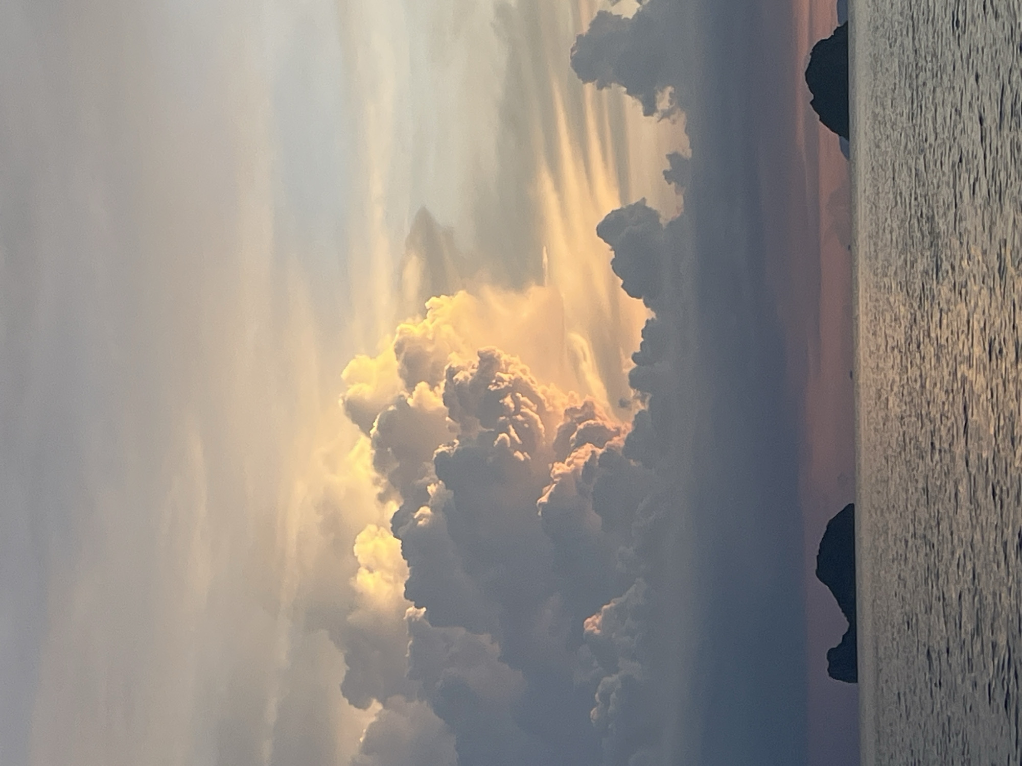 The image depicts a serene seascape at sunset near Baan Pang Ka, Thailand. The sky is filled with dramatic, voluminous clouds, some illuminated with a golden-orange hue from the setting sun, while others are in shadow, appearing a soft gray. Below the clouds, the calm sea stretches to the horizon, reflecting the muted colors of the sky. Two small, dark islands rise from the water in the foreground. The left island is larger and has a distinctive, almost horseshoe shape. The right island is smaller and more rounded. The overall atmosphere is tranquil and picturesque, characteristic of a tropical evening. There are no people or specific man-made structures visible in the scene, other than what might be distant, indistinct boats on the water.