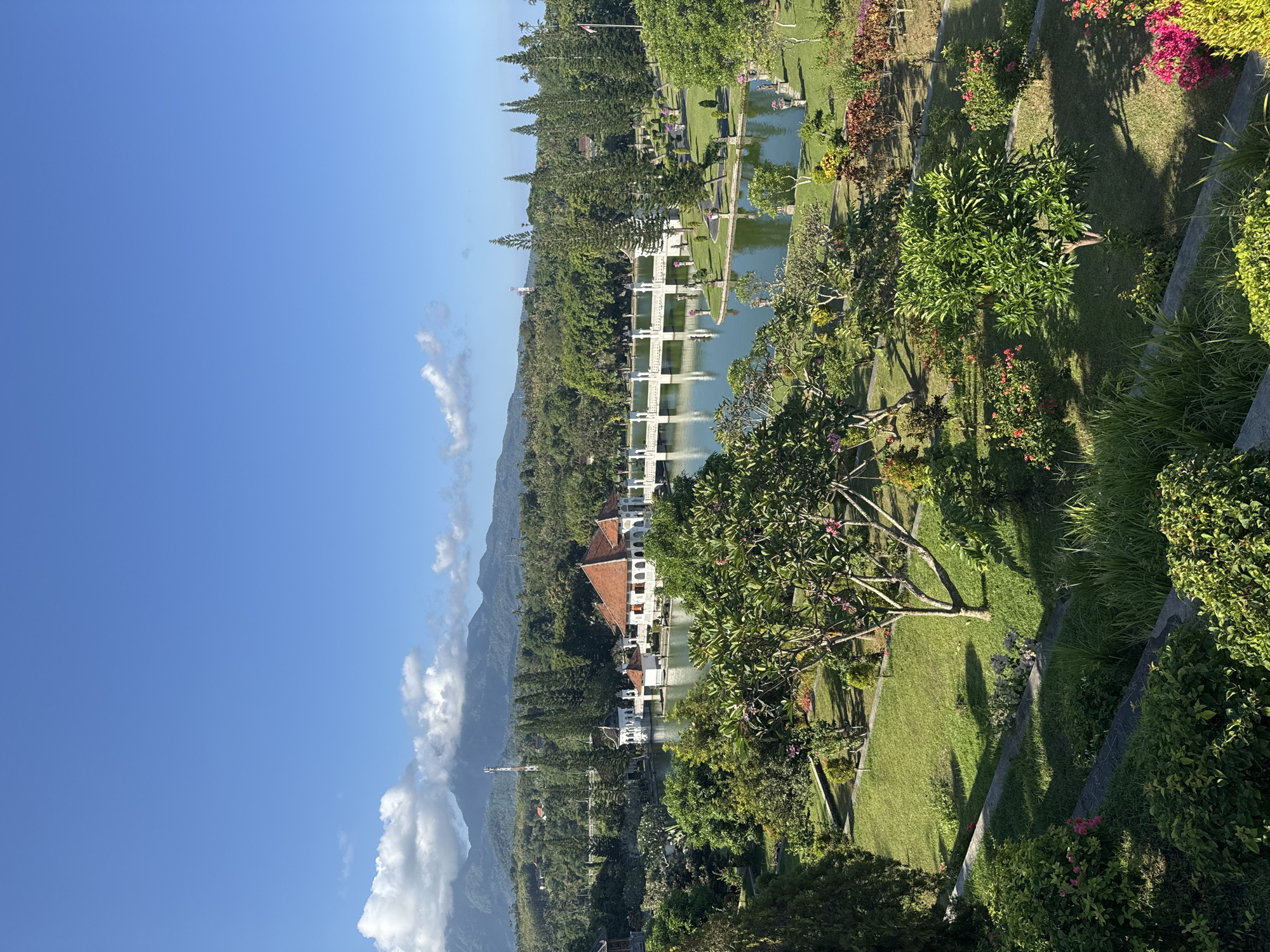 The image presents a panoramic view of a lush, expansive garden or water palace under a clear blue sky. In the foreground and midground, manicured green lawns are interspersed with a rich variety of trees, shrubs, and flowering plants, exhibiting shades of green, pink, red, and yellow. Shadows cast by trees stretch across the grass, indicating bright sunlight.
A prominent feature in the midground is a tranquil body of water, possibly a large pond or canal, traversed by several ornate white bridges or walkways. Along the edges of the water and nestled among the greenery are traditional Balinese-style buildings with distinctive reddish-brown tiled roofs and white walls. Some of these structures appear to be pavilions built directly over the water.
In the background, dense tropical foliage covers the lower slopes of large, dark mountains, which are partially veiled by a few white, fluffy clouds. The sky above is a vibrant, clear blue with scattered white clouds. The overall scene conveys a sense of serene natural beauty and architectural elegance, suggesting a well-maintained historical site or tourist attraction. The bright illumination and shadow direction indicate it is either morning or late afternoon. There are no people or visible text in the image. The location context of Karangasem, Indonesia, suggests this is likely a view of a water palace such as Tirta Gangga or Taman Ujung.