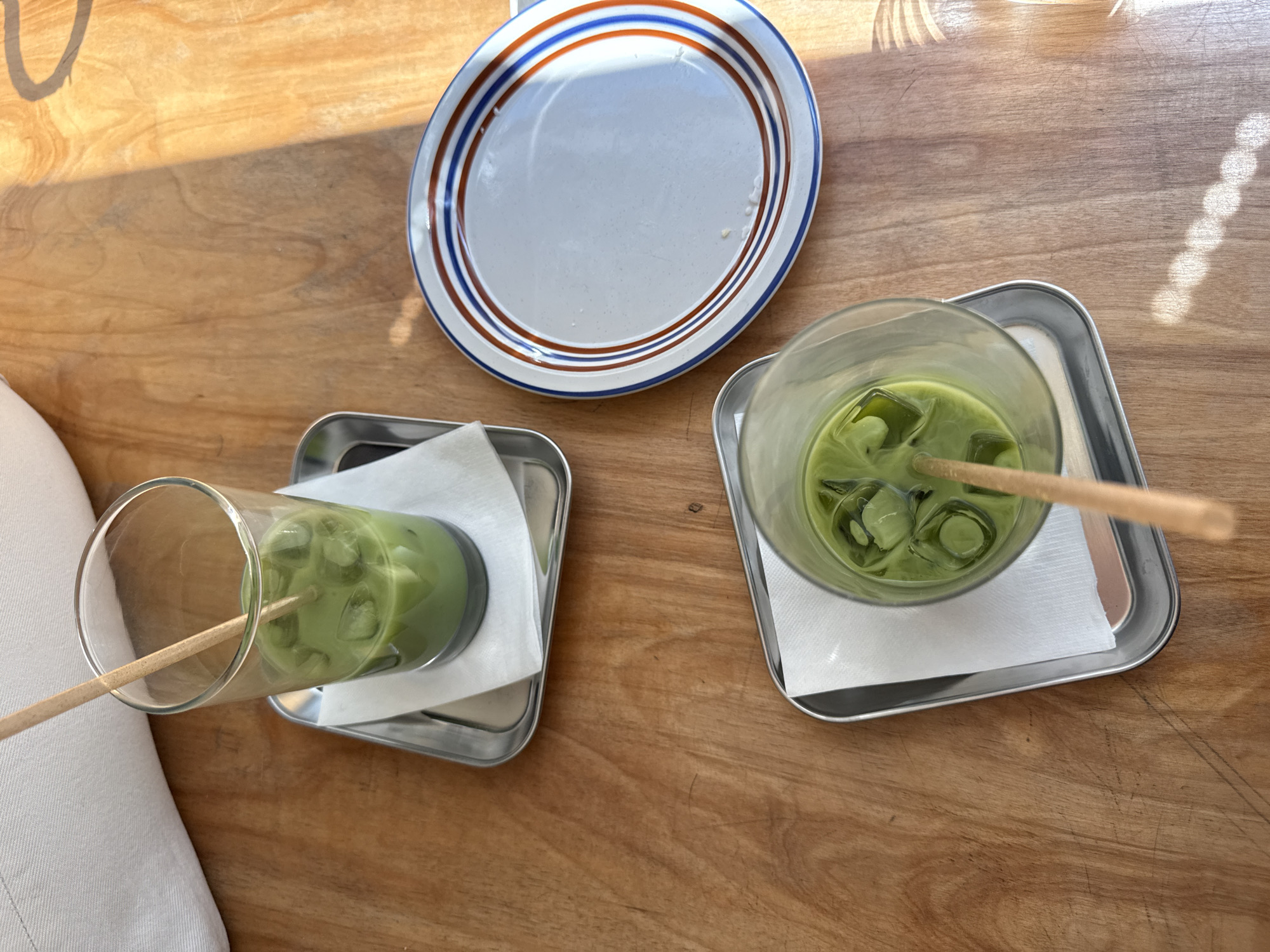 The image shows a wooden table setting with two drinks and a plate. The drinks are in tall, clear glasses, filled with a green liquid (likely matcha), ice, and a straw. Each glass sits on a small square silver tray with a white napkin underneath. Next to the drinks is an empty white plate with blue and brown stripes around the rim. The setting appears to be outdoors, based on the natural light and shadows. While the location in the city of New York, United States of America can not be determined from the information present in this image, it can be assessed that it is likely a café or restaurant setting.