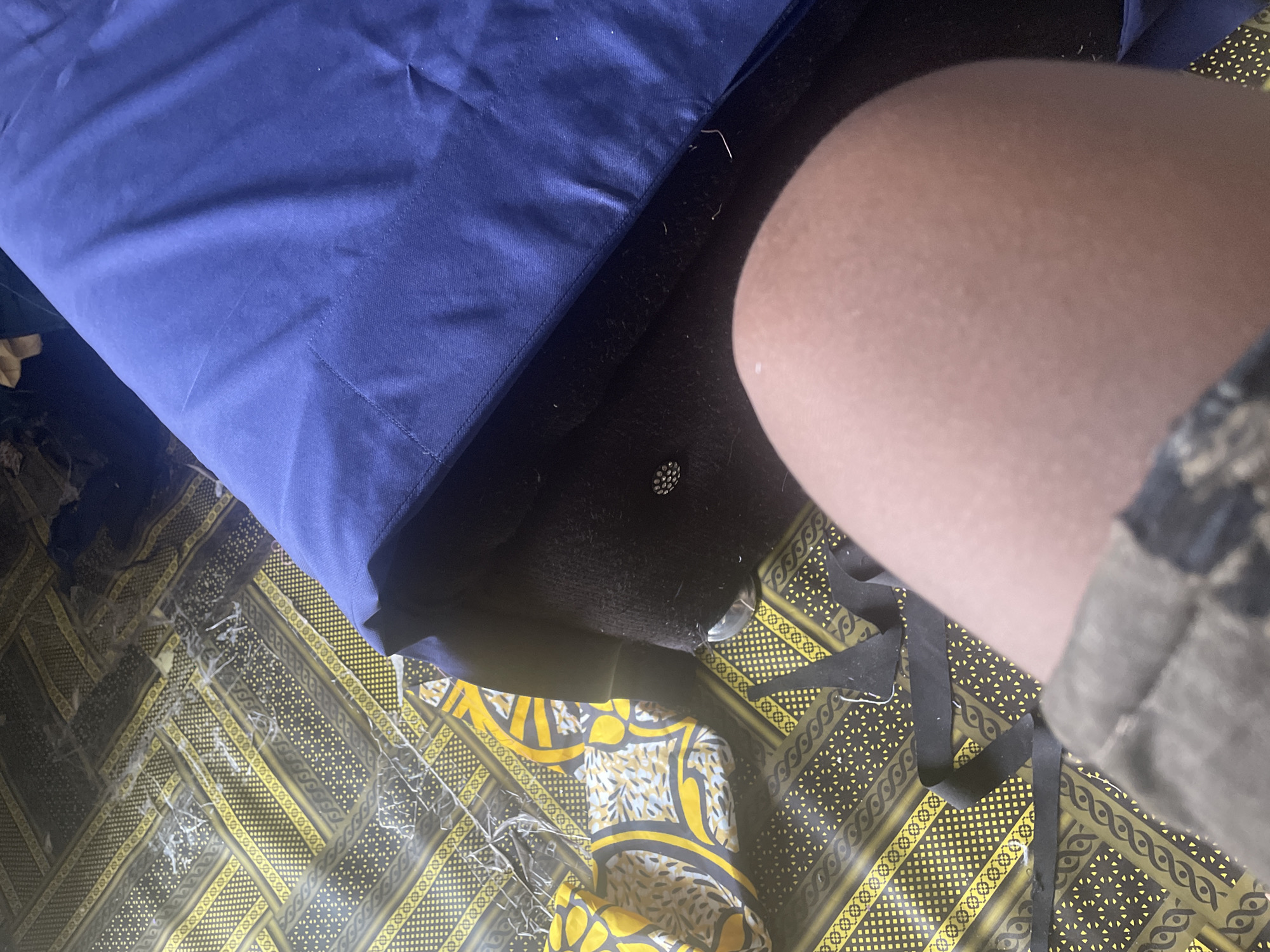 The image shows a close-up view of a person's knee and leg, with their knee positioned prominently in the foreground. The person is wearing camouflage-patterned shorts or pants. The background is a floor covered with a patterned fabric, predominantly black and yellow with intricate designs. A dark blue duvet or blanket is partially visible, draped over what appears to be furniture. There are also some loose threads and debris on the patterned fabric. The scene suggests an indoor setting, possibly a room in Jauro Yinu, Nigeria.