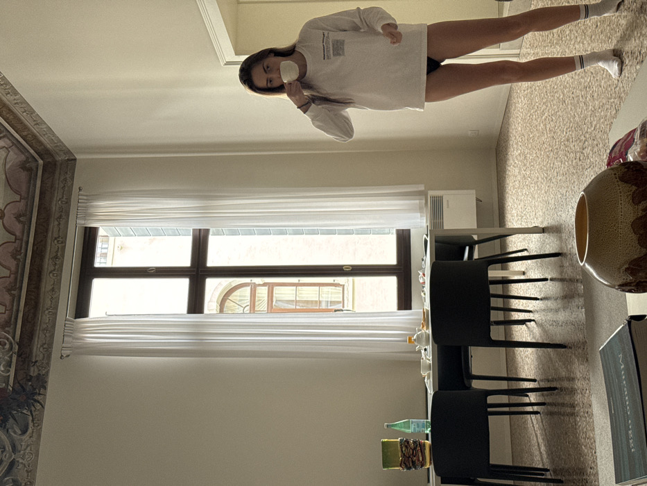 An interior shot depicts a young adult female standing in a room within a building in Venice, Italy. The room features light-colored walls, a multi-colored terrazzo floor, and a high ceiling with ornate painted detailing. A tall, multi-paned dark wooden window, framed by white sheer curtains, occupies the left side of the frame, revealing a partial view of an adjacent light-colored building with an arched window, consistent with Venetian architecture.
The female subject, with long brown hair, wears a white long-sleeved shirt with black text on the left chest, black briefs, and white ankle socks with two black stripes. She holds a white cup to her mouth with her right hand.
To her left, a white rectangular table is positioned under the window. Three black chairs are visible around it. On the table, objects include a green decorative piece, a green water bottle, a white teapot, a white cup, and plates containing residual food. A white radiator is mounted on the wall beneath the window, beside the table. In the lower foreground, a textured brown and white decorative vase and a book with the visible text "MURANO VEZZI" on its spine are situated on a surface.