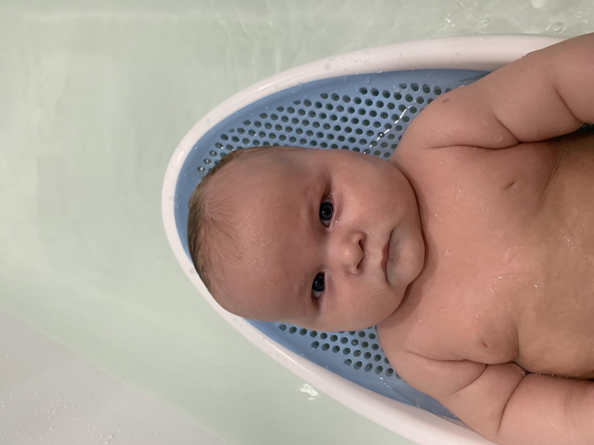 An infant is positioned in a blue and white baby bathtub insert filled with water. The baby is looking directly at the camera with a serious expression. The bathtub is in a larger tub of water, which appears to be in a bathroom. The environment is clean and well-lit. There is no visible indication of the location being in Stavropol, Russia.