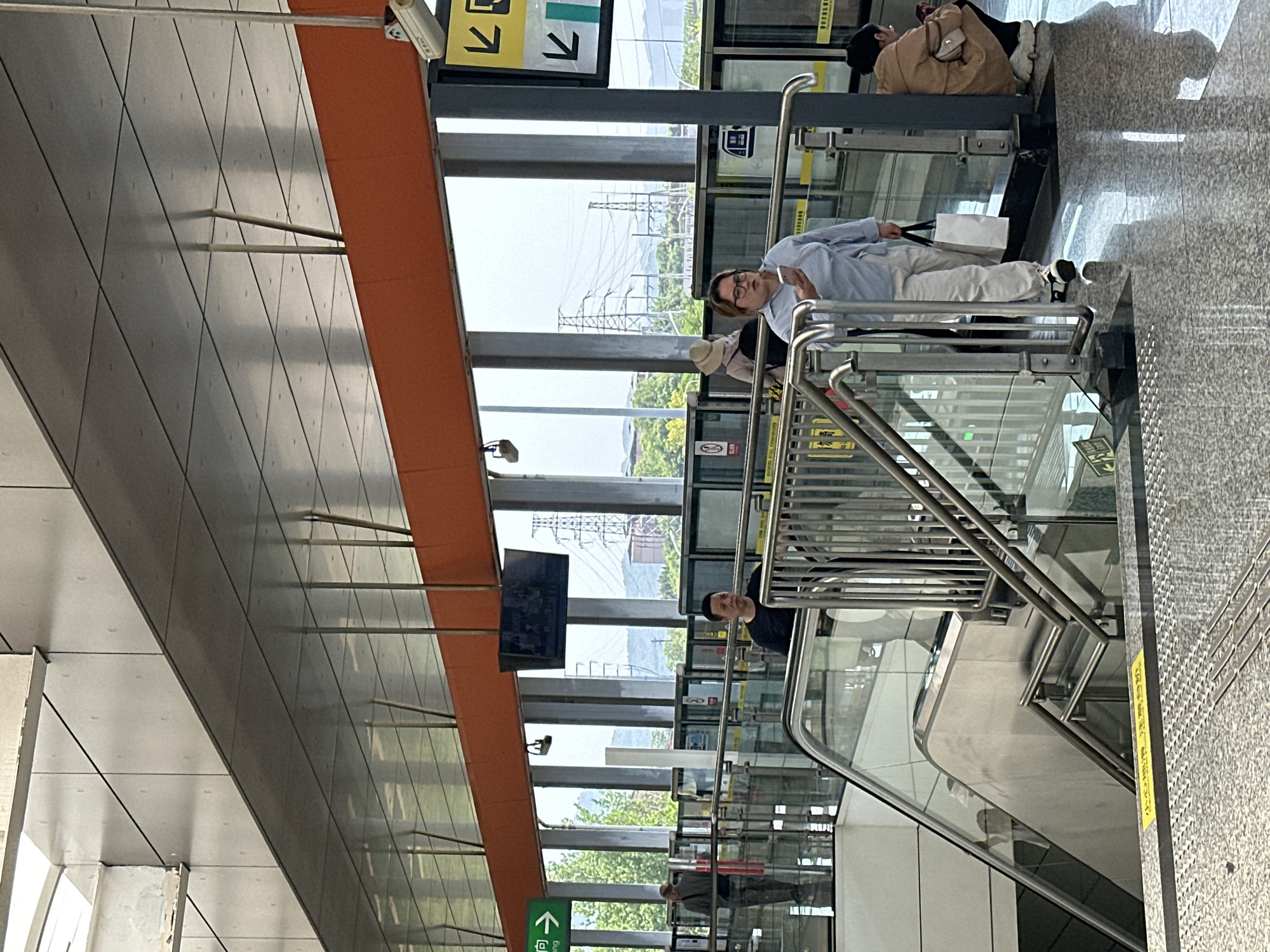 The image depicts a scene at a subway station in Jiansheng, China. The station has modern architecture with gray paneled walls, orange accents, and stainless steel railings and glass barriers. Visible through the glass windows are power lines and greenery. People are present on the escalator; one person is using a mobile device. Other subjects are the directional signs and surveillance cameras.