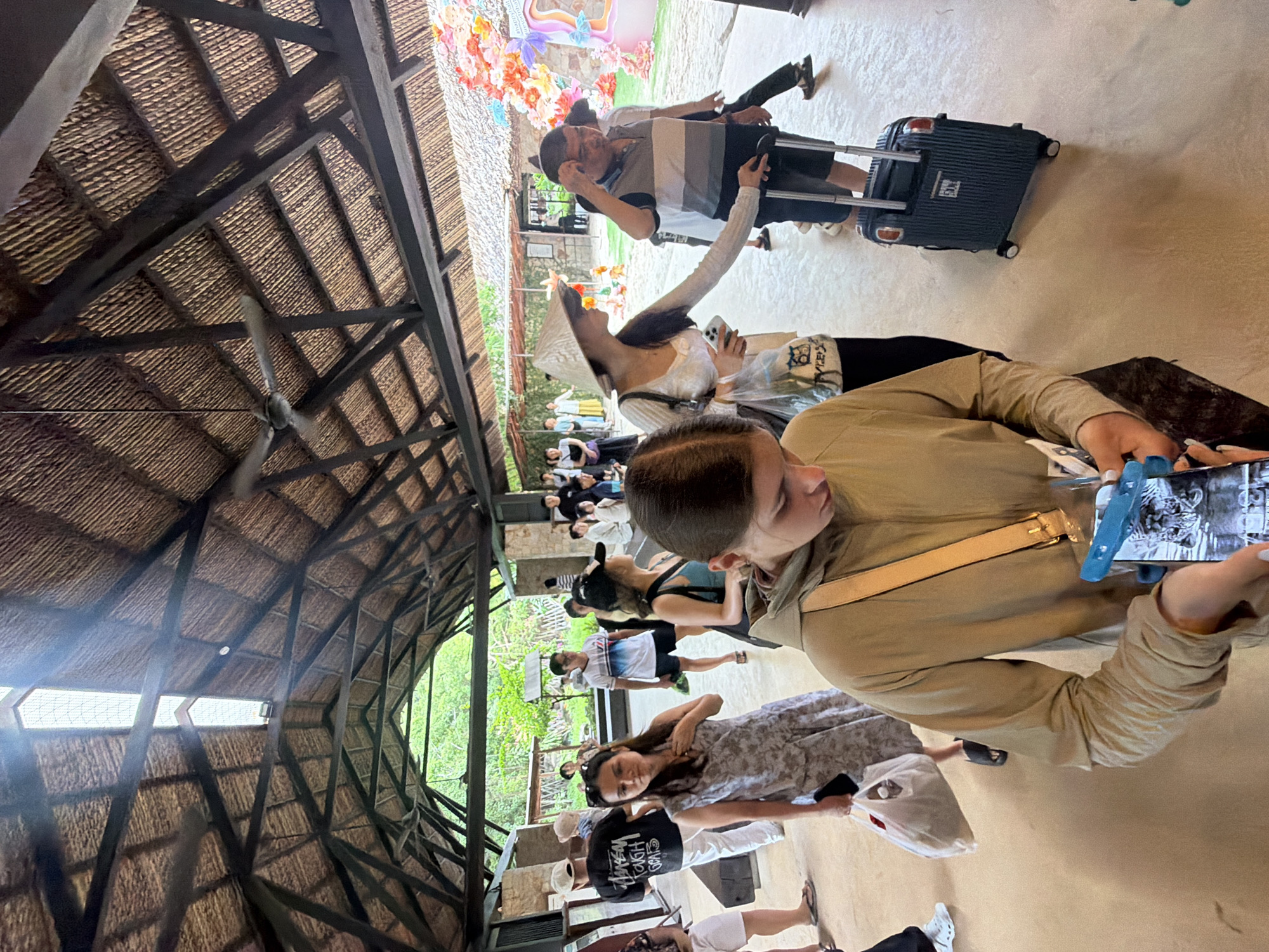 This is a medium shot of a group of people standing in what appears to be an outdoor covered walkway or waiting area. The architecture features a thatched roof supported by dark wooden beams, suggesting a tropical or rustic setting. Natural light streams in from an opening at the end of the walkway, illuminating the scene.
In the foreground, a young woman with brown hair is looking down at a smartphone she is holding. She is wearing a tan hooded jacket with a light-colored strap across her chest. To her left, a woman in a grey floral dress is also looking down, holding a white plastic bag in her left hand and a black smartphone in her right. Behind her, a man wearing a white cap and a black t-shirt with "STUSSY" printed on it stands with his back to the camera.
Further back, several other individuals are visible. A woman in a wide-brimmed conical hat and a white long-sleeved top is walking away from the camera, holding a clear plastic bag. To her right, a man in a striped t-shirt and dark shorts is standing next to a dark blue rolling suitcase, looking towards the right of the frame. Other people are seen in the background, some standing and some walking, suggesting a flow of movement or a queue.
The environment beyond the walkway appears to be green and lush, with trees and vegetation visible. There are also some colorful decorative elements, possibly floral arrangements, on the right side of the frame. The floor is a light-colored, smooth surface, possibly concrete or stone. The overall impression is of a busy area, possibly an entrance or waiting point for a tourist attraction or transportation hub. Based on the presence of tropical vegetation and the architectural style, this location could be within a resort, a cultural site, or a transport terminal in Nha Trang, Vietnam.