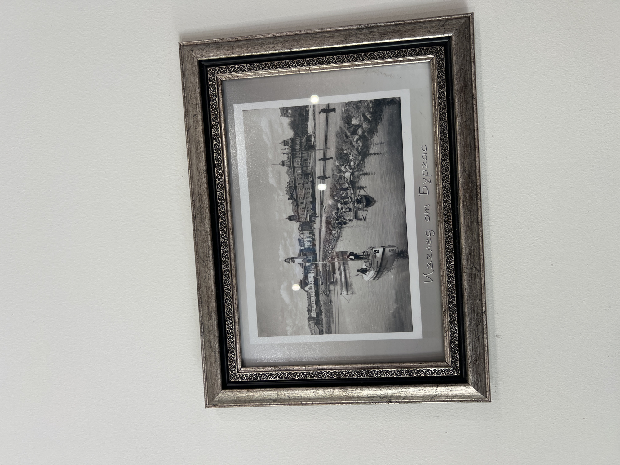 A framed black and white photograph hangs on a white wall. The frame is ornate, with a silver finish and intricate detailing. The photograph depicts a waterside scene, likely a city view with buildings along a river or harbor. The writing on the picture frame translates to "View from Budva". The image has a vintage or historical feel. There is some glare from the light on the picture.