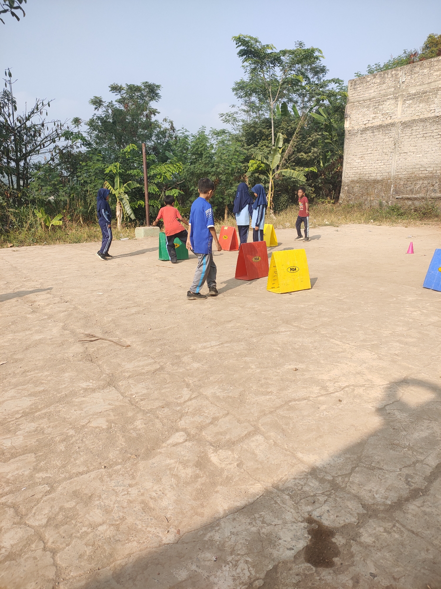 In Subang, Indonesia, on a dusty, cracked concrete playground, a group of children are engaged in an outdoor activity. The scene, likely taking place on a sunny day, features several children scattered across the area. One boy, wearing a blue shirt and grey pants, is in the foreground, his body angled as if mid-movement. Another boy in a red shirt stands near a green cone in the background. Two children in blue hijabs and matching outfits are also present, positioned to the left of the boy in red. A small child in a red shirt and dark pants is further in the background, facing away from the viewer. Several brightly colored cones, red, yellow, and blue, are strategically placed around the playground, some marked with the acronym "POA." The background is a mix of lush green trees, a banana tree, and a weathered, light-colored brick building. The sky is a clear, pale blue. The overall impression is one of a casual outdoor educational or recreational event for children.