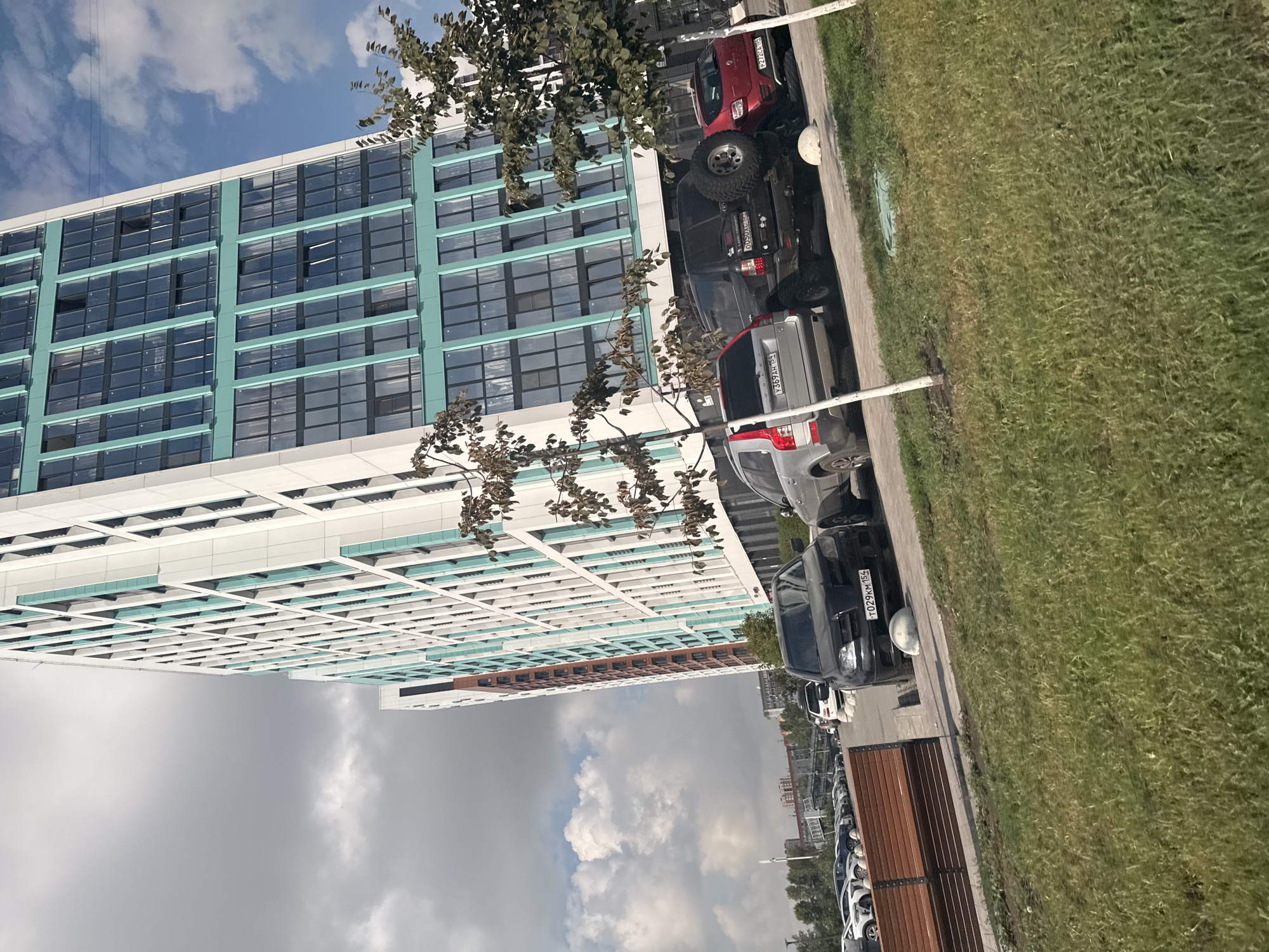 A street-level, eye-level, medium shot captures a view of a modern, tall residential building with a facade of white and teal panels, interspersed with dark-tinted windows. The building stands at an angle to the camera, suggesting a perspective from a street or open area. In the foreground, a patch of green grass slopes gently upwards, with a wooden bench visible to the left. The middle ground is occupied by a parking lot with several cars parked.
The cars visible are a black sedan with a license plate reading "T029KM 154", a black SUV with a spare tire mounted on the rear and a license plate reading "0050XC 154", and a red hatchback with a license plate reading "P212EA 154". Behind these, a silver SUV is partially visible. A white spherical bollard is also present in the parking lot.
In the background, a row of parked cars stretches along the street, and further back, other residential buildings and a pedestrian bridge can be seen. The sky above is partly cloudy with scattered white clouds against a blue sky. The overall impression is a contemporary urban environment.
Based on the visible license plates with the "154" region code, the scene is located in the city of Novosibirsk, Russia.