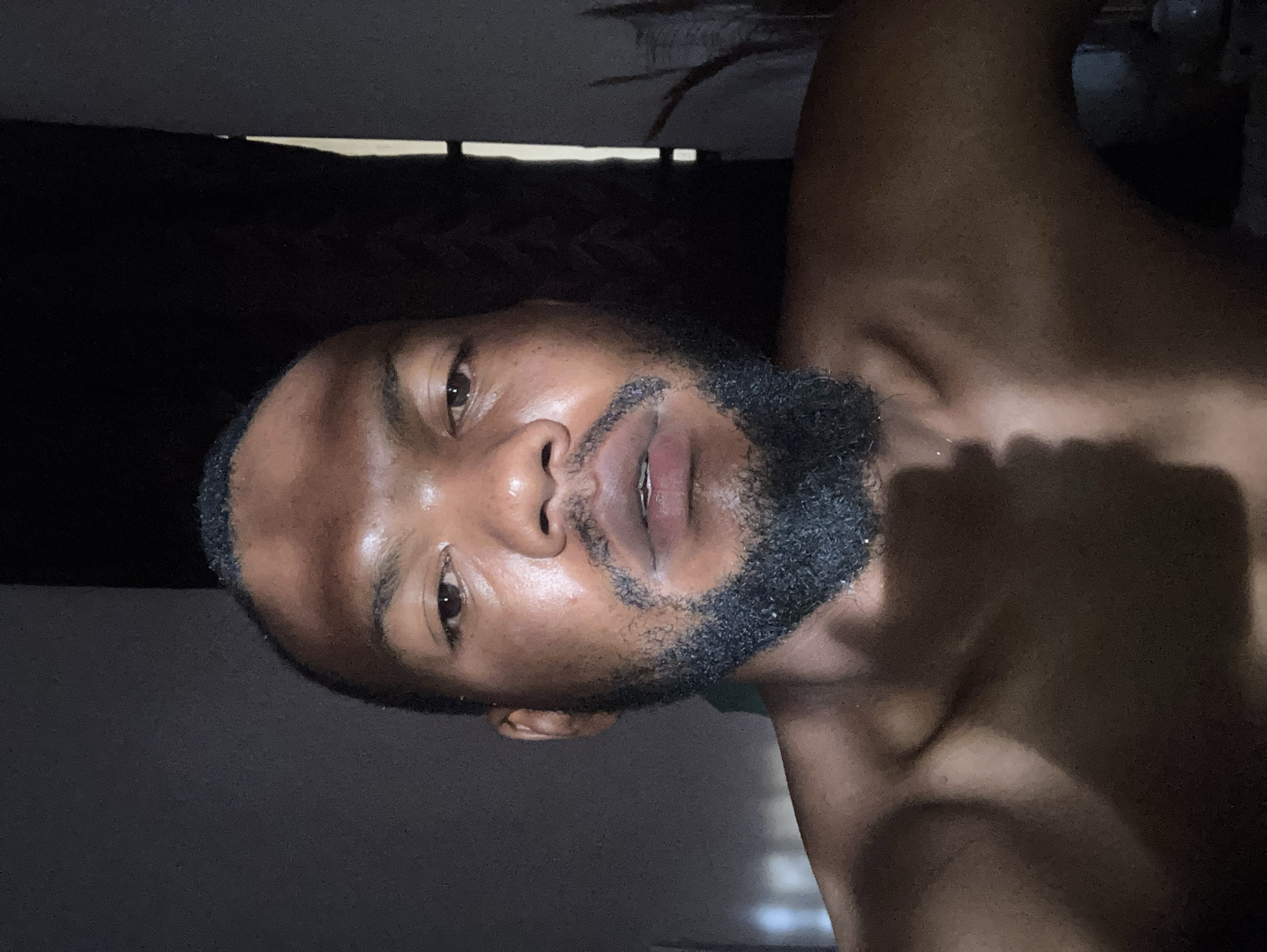 The image is a close-up, portrait-style shot of a shirtless dark-skinned man with a beard. The man's face is the primary focus, showing his eyes, nose, and mouth in detail. The lighting in the image is dim, casting shadows on his face and body. The background is dark and indistinct, with what appears to be a window or curtain visible in the upper left corner. There is no information about the location or city of Jos, Nigeria, in the image. There are no interactions or actions visible in this photograph. The environment is dark and indistinct.