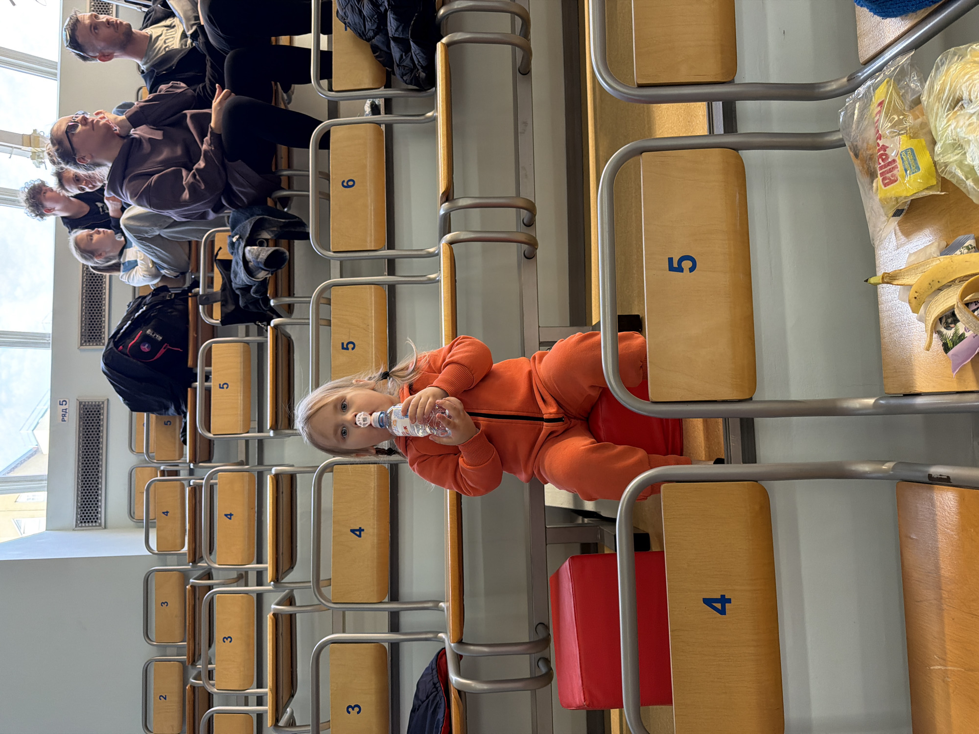 The image depicts a young child in an orange jumpsuit, seated on a red cushion on a bench, and drinking from a plastic water bottle. The bench is part of a row of stadium-style seating, numbered "3" through "6". Adults are sitting on benches behind the child. There are bags and other items on the benches around the seated individuals. The photo appears to be taken inside a building with a white wall.