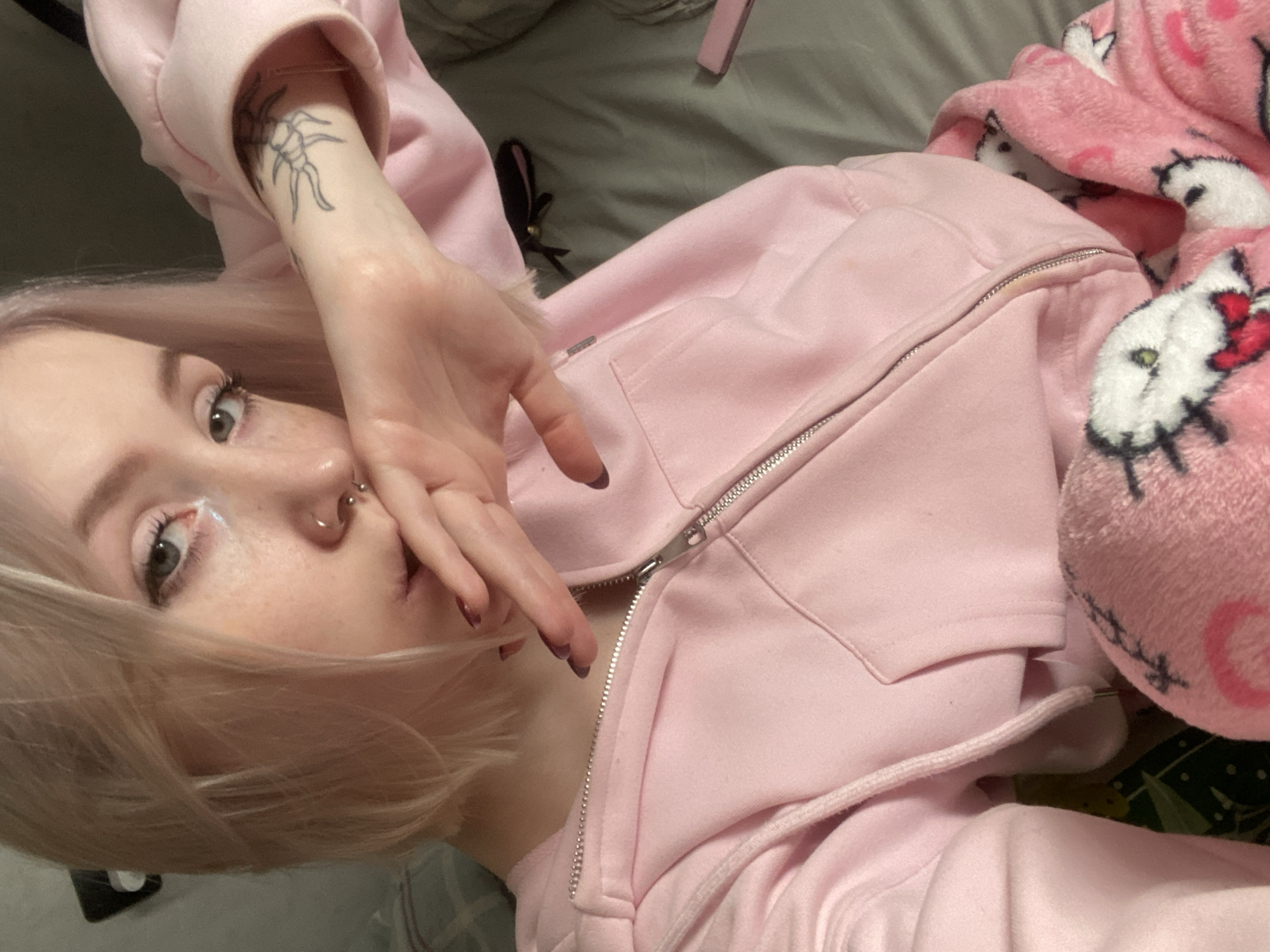 The image, taken in Izhevsk, Russia, captures a close-up, angled view of an individual lying down indoors, likely in a bedroom setting. The person, positioned in the lower-left portion of the frame, has light blonde hair, pale skin, and visible freckles across their nose and cheeks. Their light-colored eyes, possibly grey or green, are directed towards the viewer. They wear makeup including dark mascara and a bright, possibly white, eyeliner or highlight at the inner corner of the eye. A septum piercing adorns their nose.
The individual is wearing a light pink zip-up hoodie, which is partially unzipped. Their right hand, featuring dark-colored nail polish, is raised and covers part of their mouth and nose. A linear tattoo, resembling an insect or arachnid with multiple legs, is visible on their inner left forearm, partially obscured by the hoodie sleeve.
To the right of the person, a soft, pink blanket patterned with multiple Hello Kitty faces is prominent. Each Hello Kitty character has its signature red bow and visible eyes (green or yellow). The bedding beneath the person appears to be a plain grey or olive-green fabric. Faintly visible in the background are a small pink object, possibly a phone case, and a dark accessory with a small bell. The person's expression is neutral, and their pose suggests a moment of rest or casual interaction. There is no discernible text within the image.