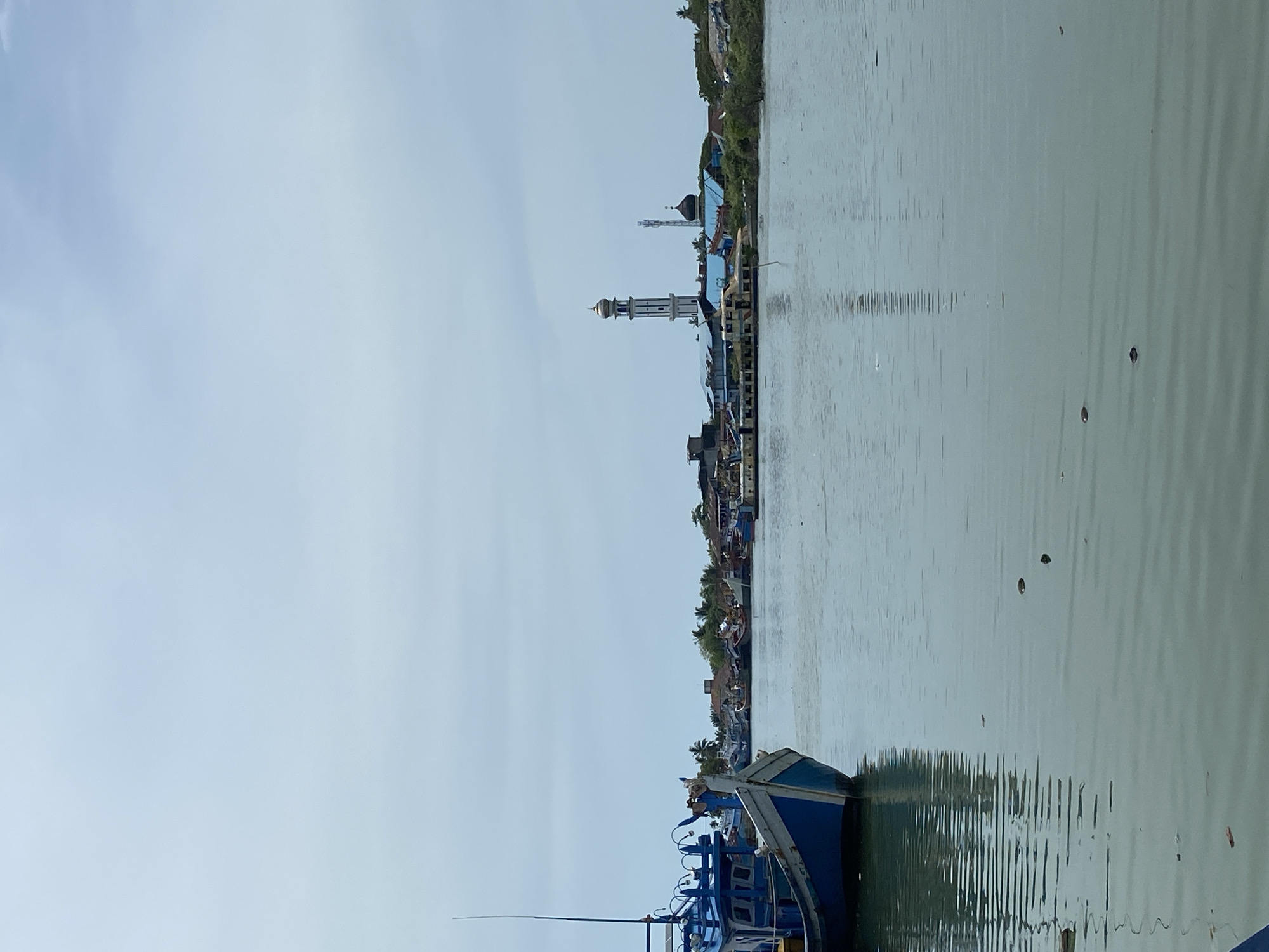 A sideways medium shot shows a view of Tanjongkapai, Indonesia, across a body of water, from the perspective of a boat.
The foreground features the bow of a blue and white boat with a visible antenna. The water is calm, with slight ripples and reflections of the cityscape.
Across the water, the cityscape includes buildings, houses, and greenery along the shoreline. Prominent features include a white mosque with a tall minaret topped with a golden dome, and another minaret in the distance. The sky is overcast.