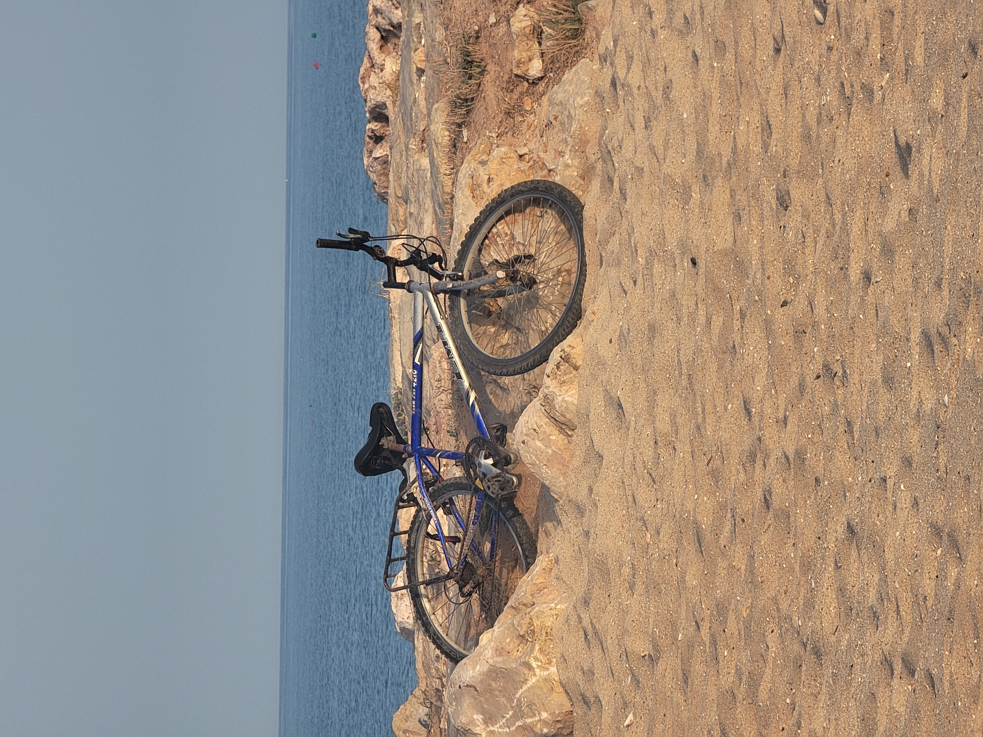 A blue and silver mountain bike is resting on a rocky shore near the sea. The bicycle is positioned diagonally, with its front wheel facing the right side of the frame. The sand in the foreground is coarse and shows signs of being disturbed, possibly by footprints. The background consists of calm blue water stretching to the horizon, under a pale, cloudless sky. Several buoys are visible in the water, indicating a marked area. The rocks on the shore are rough and uneven, with some sparse vegetation growing between them. The overall scene suggests a peaceful, natural environment.