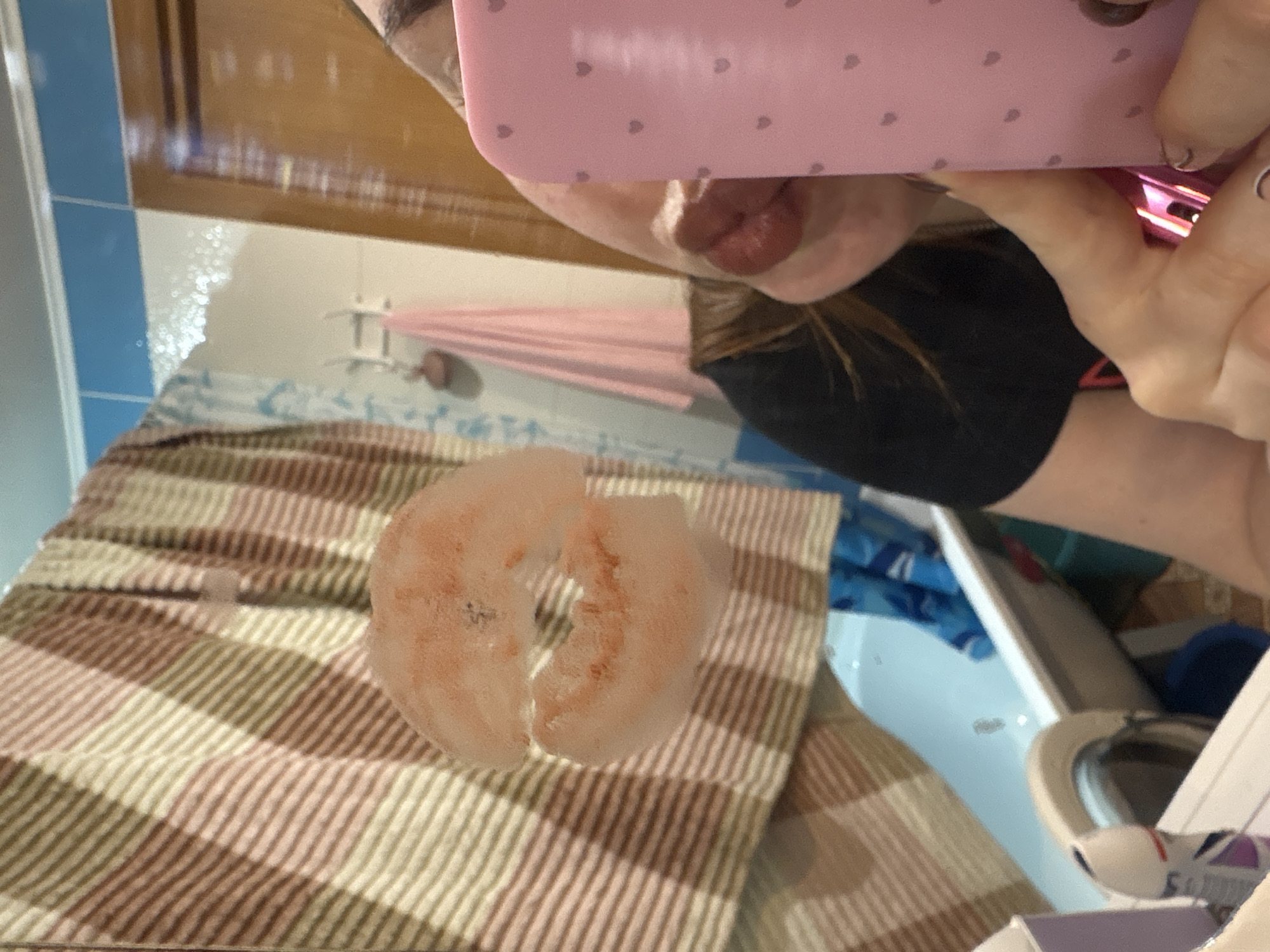 A selfie-style image shows a person partially visible behind a pink phone, taking a picture of a thawed shrimp ring on a striped towel in what appears to be a bathroom. The background includes a shower curtain, blue tiled walls, and other bathroom items. No specific location in Izhevsk, Russia, is discernible from the image.