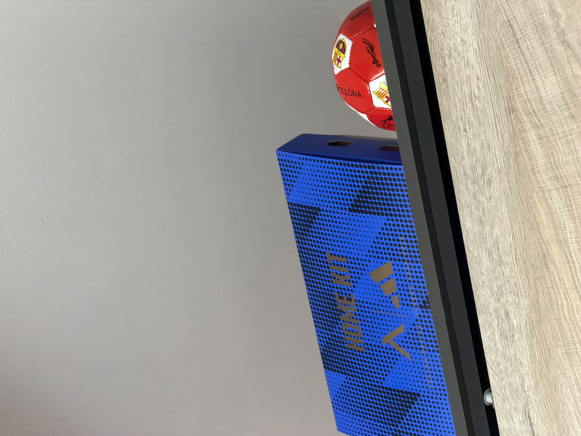 The image shows a close-up view of a shelf with a blue Adidas box and a red and yellow soccer ball. The box is decorated with a black dotted pattern and the words "HOME KIT" and the Adidas logo are visible in gold. The soccer ball appears to be a Barcelona F.C. ball. The background is a plain, light gray wall. The image does not provide any information about a specific location in the city of Roman, Bulgaria.