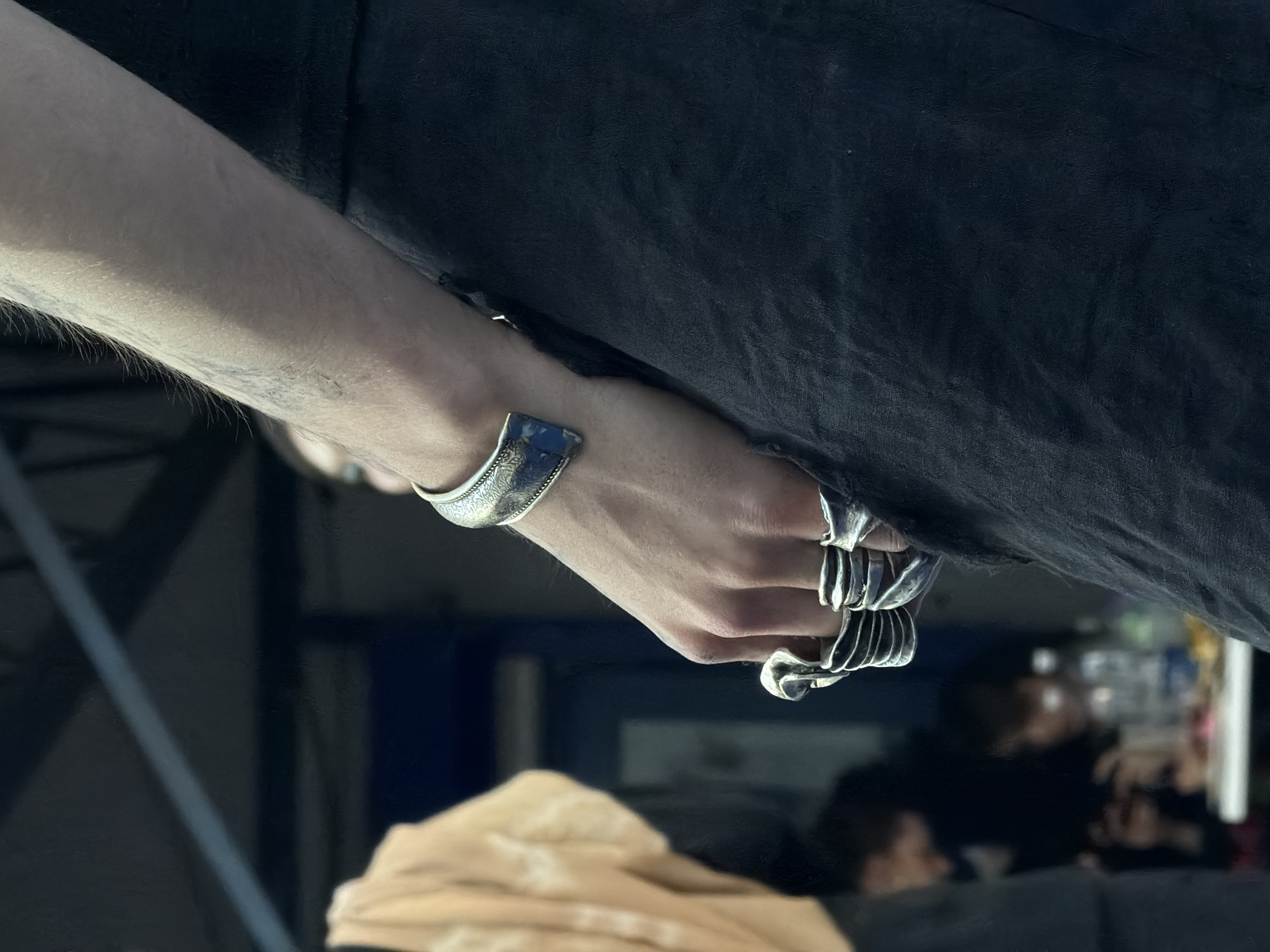 A close-up shot depicts a light-skinned arm and hand adorned with silver jewelry, specifically a bracelet and multiple rings. The hand rests against a backdrop of dark fabric or clothing, likely leather, with its edge visible near the hand. The background is somewhat blurry but reveals a scene with a pale beige or light-yellow object at the bottom, possibly fabric or furniture. Further in the background, there appear to be human figures or reflections, possibly indicating an interior space. The location is identified as Bishkek City, Kyrgyzstan. No specific actions or interactions are directly observable.