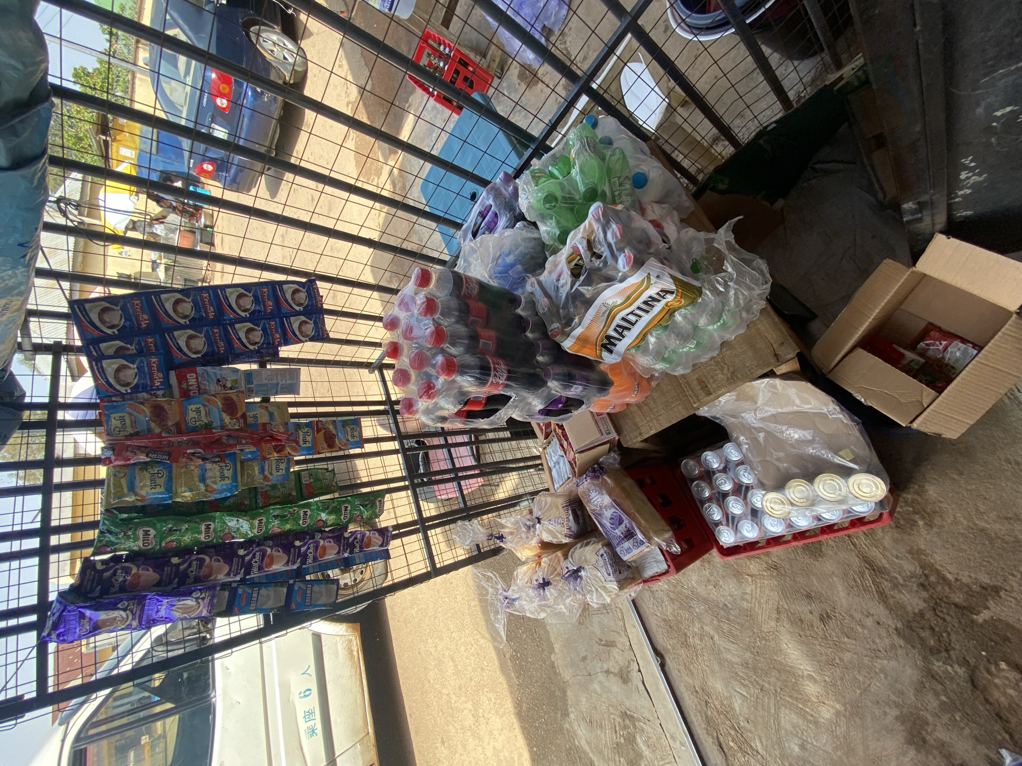 This image captures an outdoor street market stall in Gura Top, Nigeria, displaying a variety of beverages and packaged goods. The stall is a metal cage structure, with items neatly arranged for sale. In the foreground, several loaves of bread are visible, some individually wrapped in clear plastic and others in larger plastic bags. A red crate holds canned drinks, some still sealed in plastic wrap. Stacked bottles of soda, including Coca-Cola and what appears to be Maltina, are also prominent. Smaller packets of beverage powders, such as Milo and Hot Chocolate, are hanging from the metal bars of the stall. In the background, a white van with Japanese characters ("乗車 6人" meaning "6 people boarding") is parked to the left. A blue car is visible further back, and a small yellow structure with a white umbrella suggests other market activity. The ground is paved with a rough, brownish material. Sunlight casts shadows, indicating a daytime scene. No individuals are clearly identifiable within the frame, suggesting the stall is either unattended or the proprietor is not in the immediate vicinity of the camera's focus.