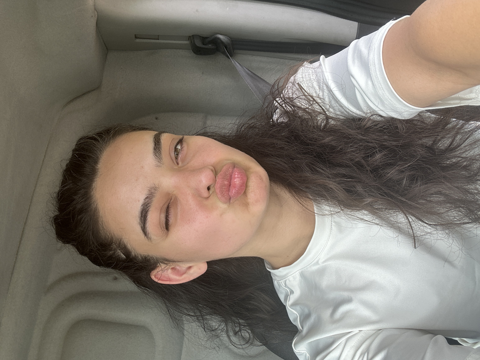 A young woman with long, dark, curly hair is taking a selfie in a vehicle. She is wearing a white long-sleeved t-shirt. Her right eye is closed, and her left eye is open, looking at the camera. Her lips are pursed as if she is about to kiss. The background of the image shows the interior of a vehicle, with a gray fabric lining the walls and a seatbelt visible on the right side. The lighting suggests it is daytime. There is no visible indication of the location being in Malevo, Bulgaria.