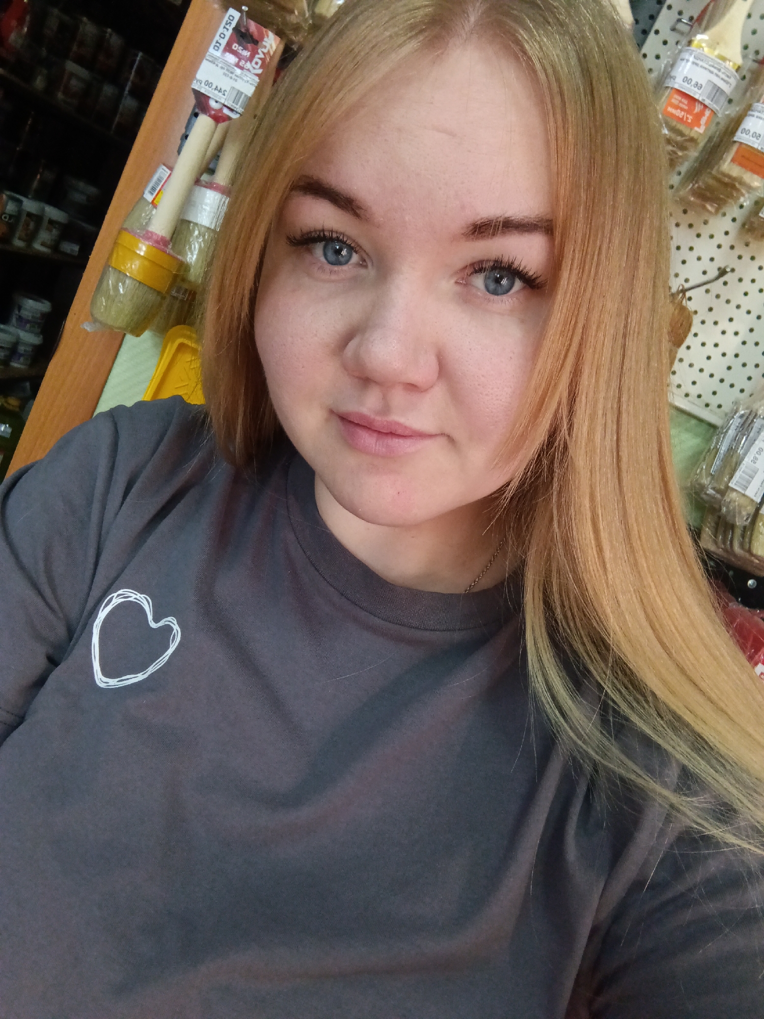 A young, fair-skinned woman with light blue eyes and strawberry blonde hair is taking a selfie indoors. She is wearing a charcoal gray t-shirt with a small, white heart design on the left side. She is positioned in front of a retail display of paintbrushes, paint cans, and other hardware supplies. The location is a store in the city of Фоки, Russia.