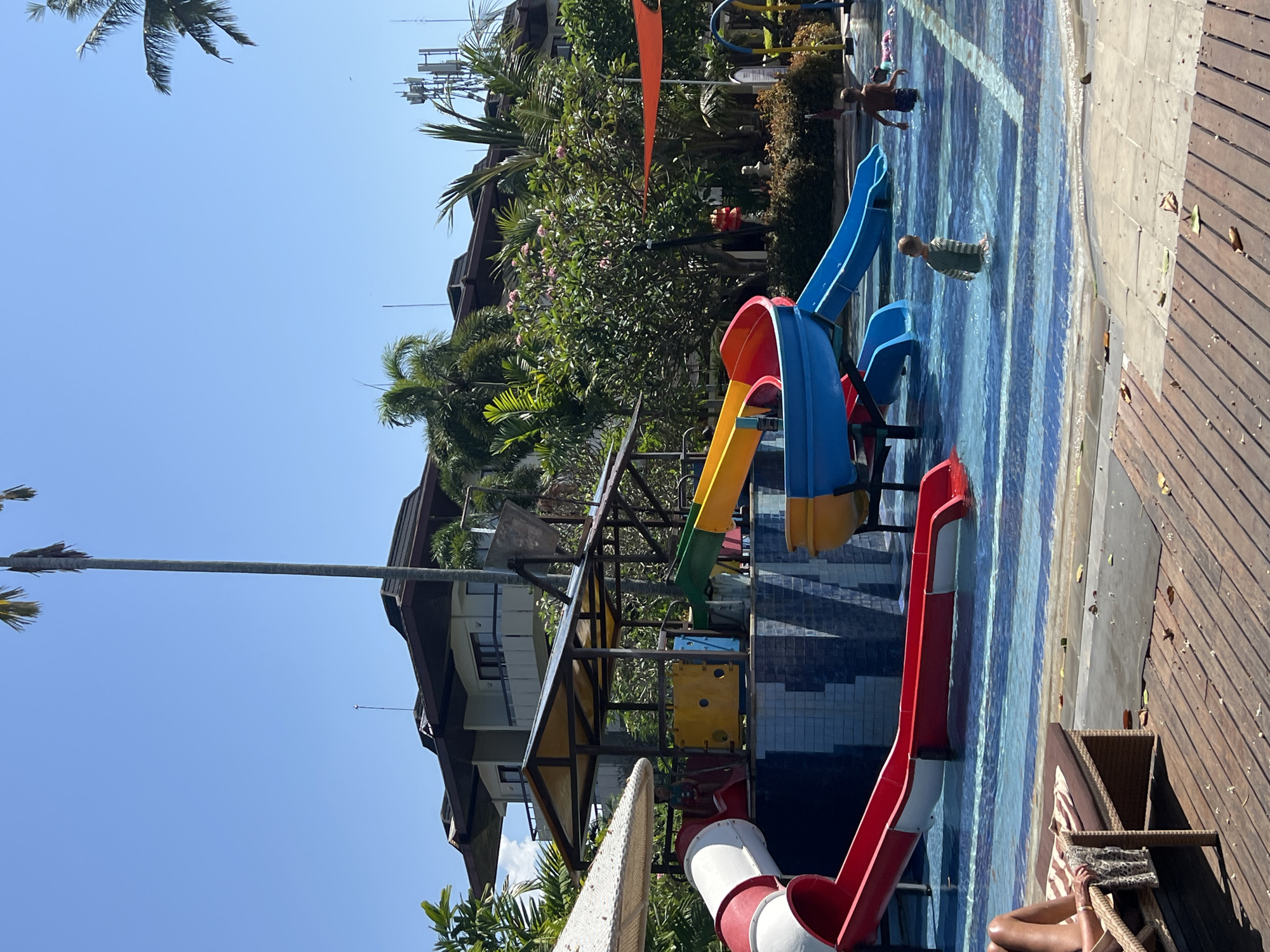 In Denpasar, Indonesia, this outdoor swimming pool complex features several water slides, including a large red and white tube slide and a multi-colored, multi-level slide structure with yellow, blue, and red sections. A child wearing a green and white striped shirt stands in the shallow end of the pool, while another child, seen from the back, is further in the water. A person's legs are visible as they recline on a lounge chair on a wooden deck to the left of the frame. In the background, a multi-story building with balconies and dark roofing is surrounded by lush green trees, including palm trees, under a clear blue sky. There are also communication towers visible in the distance.