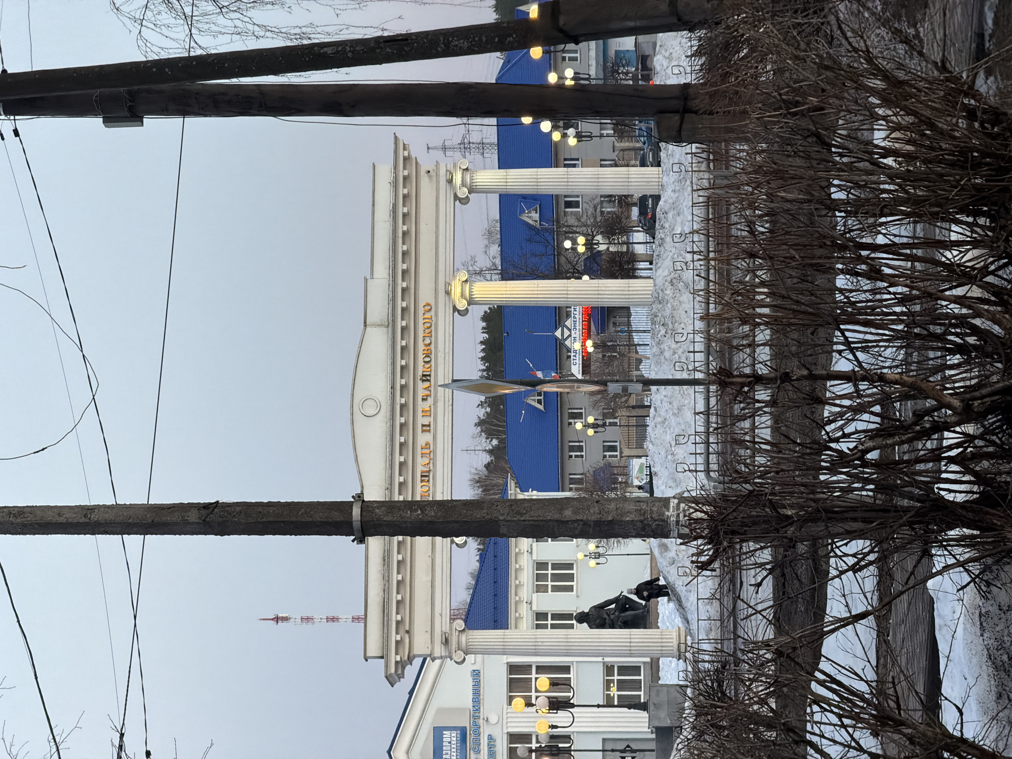 The image depicts a view of a square in the city of Chaikovsky, Russia, featuring an architectural structure with columns and Cyrillic text. The columns flank an entrance to the square, with buildings having blue roofs visible in the background. A statue is visible on the lower part of the image. Bare trees and utility poles are present in the foreground and along the sides. Traces of snow are also visible on the ground, suggesting a winter or cold-weather setting. The overcast sky contributes to the flat lighting of the scene.