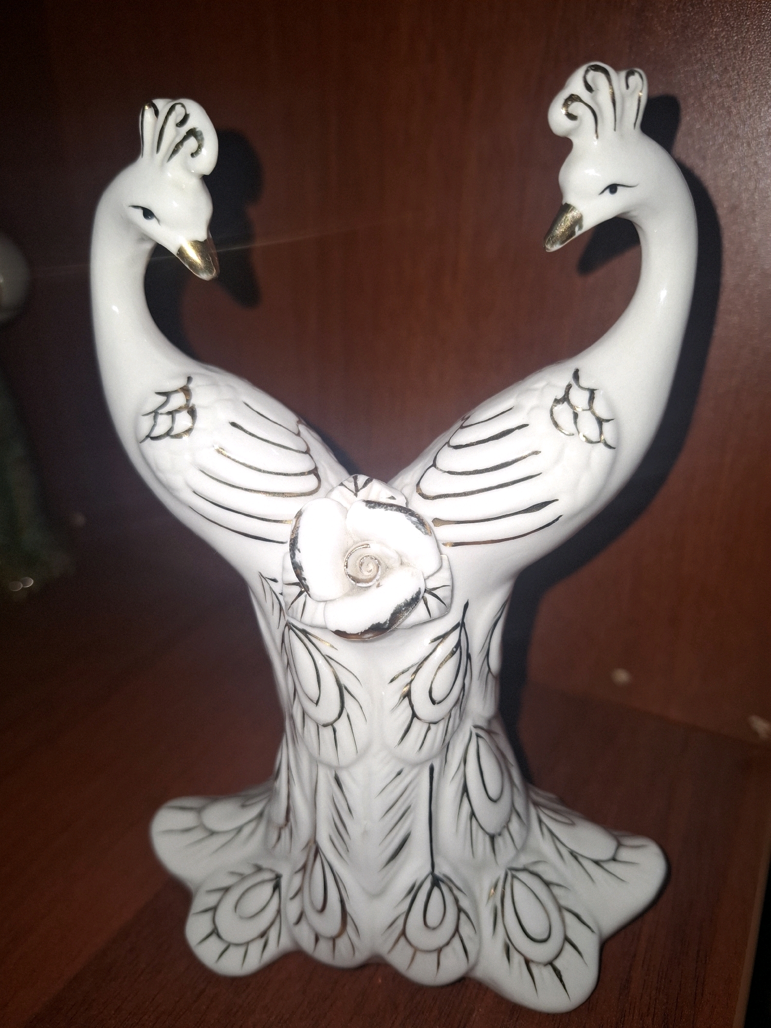 A white porcelain figurine of two peacocks facing each other is centrally located on a wooden surface, likely a shelf or table. The peacocks have gold-tipped beaks and black and gold detailing on their feathers and crests. A white rose with gold accents is positioned between the necks of the peacocks. The background is a dark brown, textured surface, suggesting a wooden cabinet or wall. The lighting is subdued, casting shadows that highlight the sculptural details of the figurine. The setting is domestic, possibly a home interior in Almaty, Kazakhstan.
