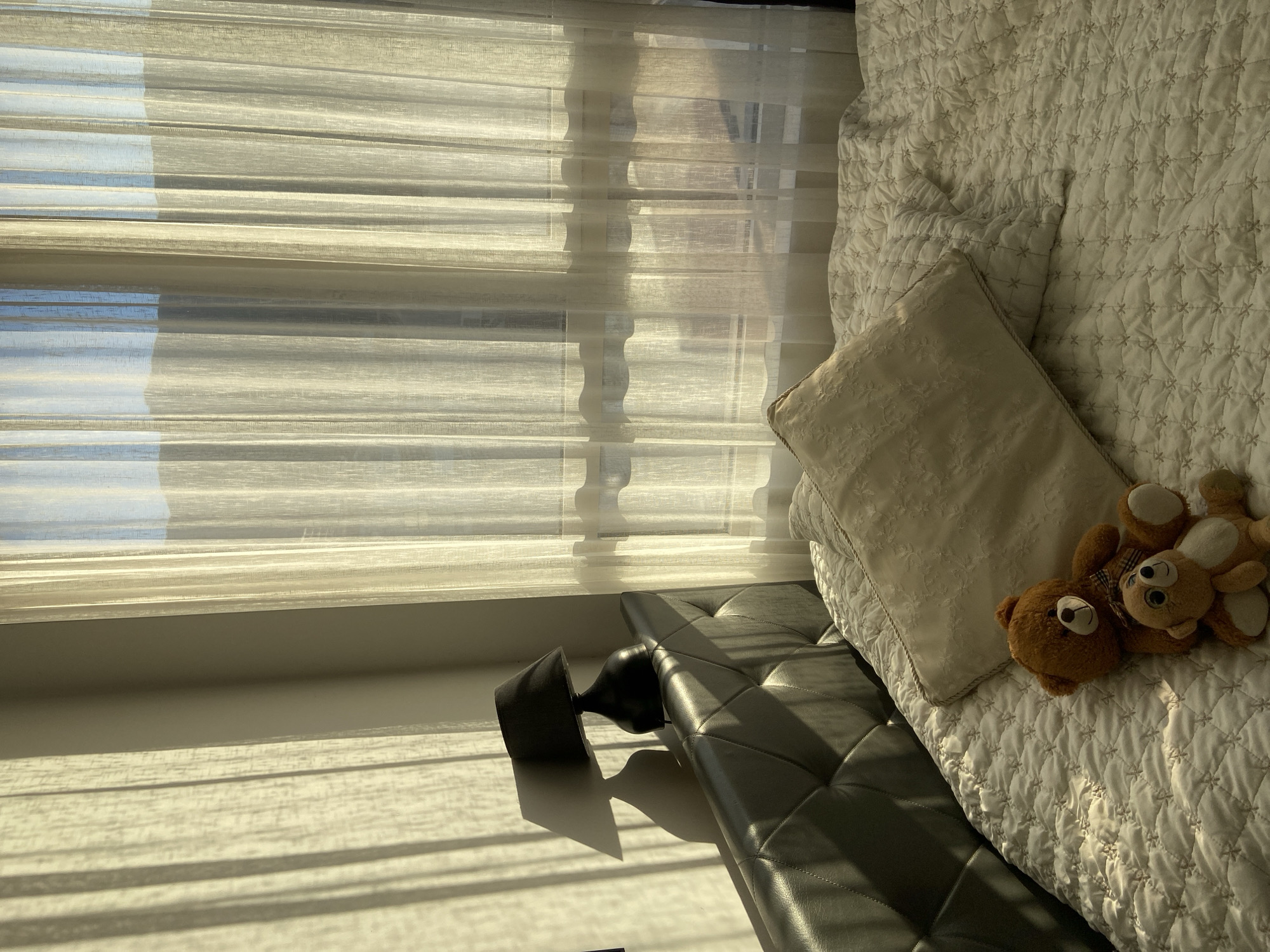 The image depicts an interior scene with strong sunlight filtering through a window covered by sheer curtains. A portion of a bed or daybed is visible on the right, covered with a white quilted blanket, a light-colored pillow, and two stuffed teddy bears. To the left and below the bed is a dark, possibly leather, bench. An ornamental piece or object is located at the edge of the bench. The location in the city of Budva, Montenegro cannot be determined based on visual information alone. No people or direct actions are visible.