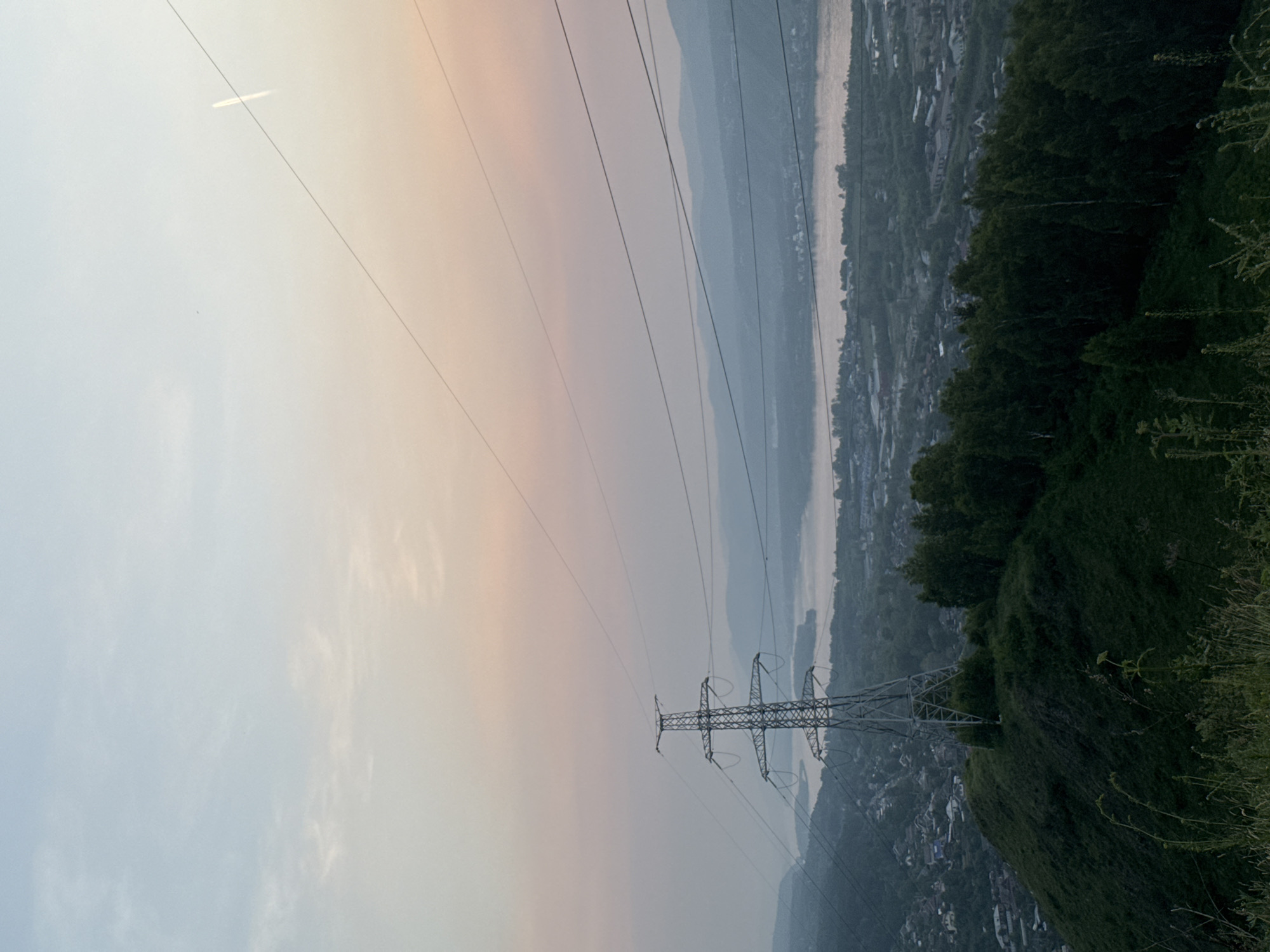 The image depicts a wide view of a valley with a river running through it, taken from a high vantage point. A towering electricity pylon dominates the foreground on the right side, with power lines stretching across the frame. The valley below is densely populated with houses and buildings, nestled amongst lush green hills and trees. The far side of the valley is lined with hazy, layered mountains. The sky transitions from a pale, bright blue on the left to a soft, peachy hue on the right, suggesting either sunrise or sunset. The overall atmosphere is serene and captures a blend of human infrastructure and natural landscape. The scene is set in Krasnoyarsk, Russia. There is no visible text in the image.