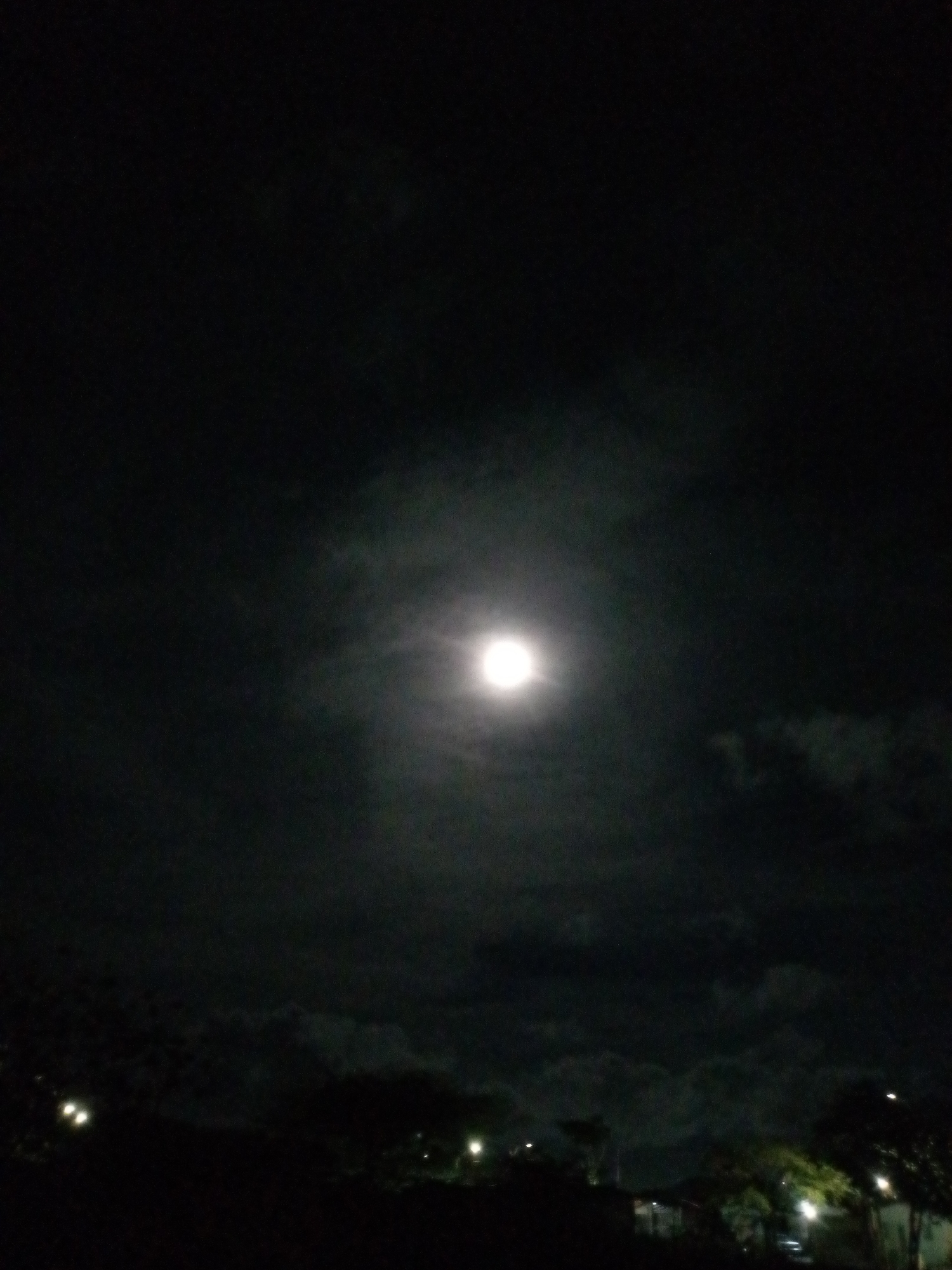 A nighttime scene depicts the moon, partially obscured by clouds, dominating the sky. In the foreground, silhouettes of trees and buildings suggest a landscape. Faint lights, potentially from street lamps or building illumination, are scattered across the lower portion of the image, adding to the ambiance. The city of Altagracia, Venezuela is not discernible in the image.