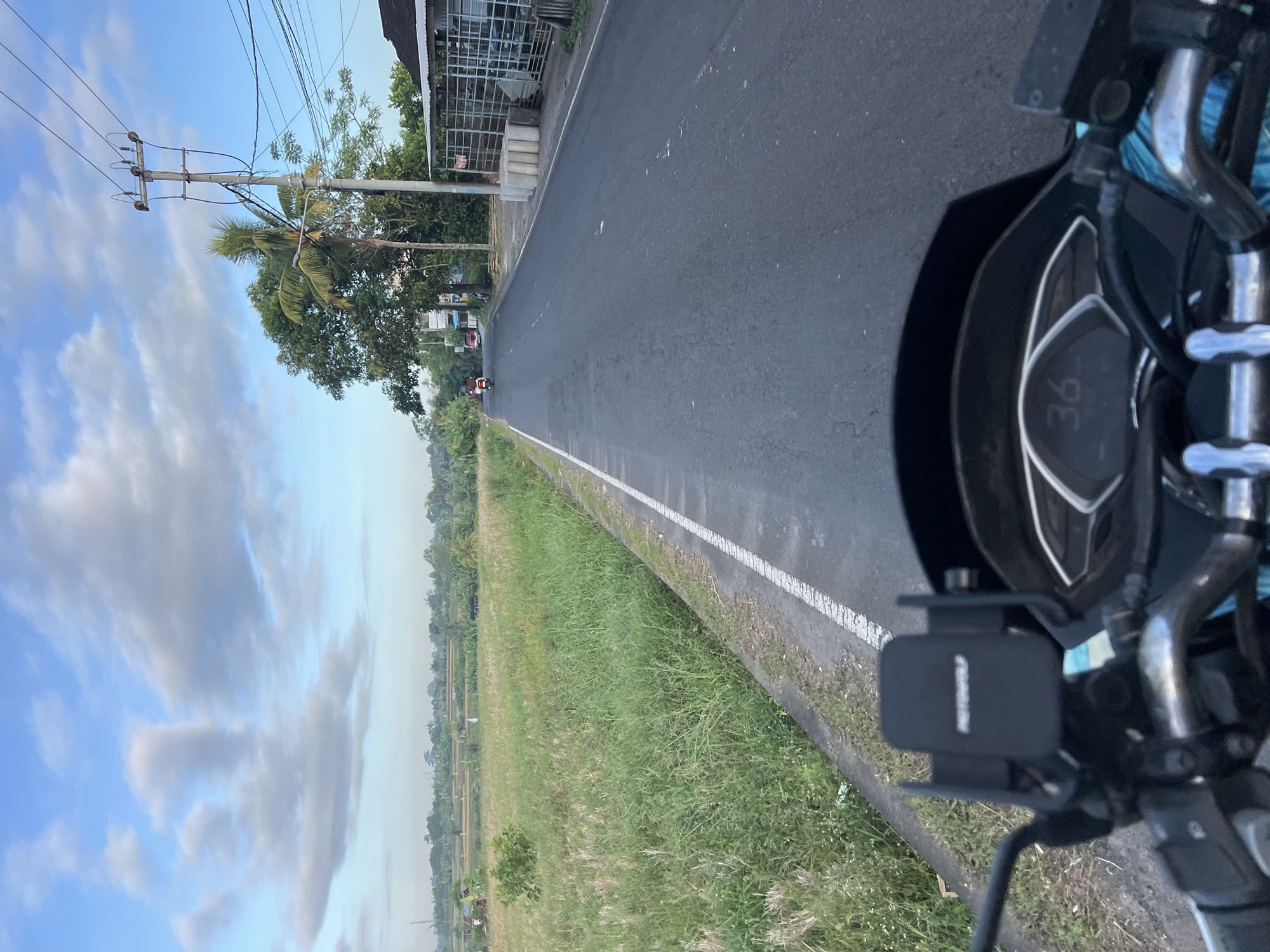 The image is a first-person perspective from a motorcycle on a paved road in Abiansemal, Indonesia. The road is lined with green fields and vegetation on the left and buildings and utility poles on the right. The sky is partly cloudy with a clear blue expanse above. In the foreground, the handlebars and dashboard of the motorcycle are visible, with a phone mount and a digital speedometer displaying "36". In the middle ground, there are a few vehicles on the road, including a scooter and a red car. The background features more greenery, palm trees, and scattered buildings, suggesting a rural or semi-rural setting. The overall scene depicts a serene journey through the Indonesian countryside.