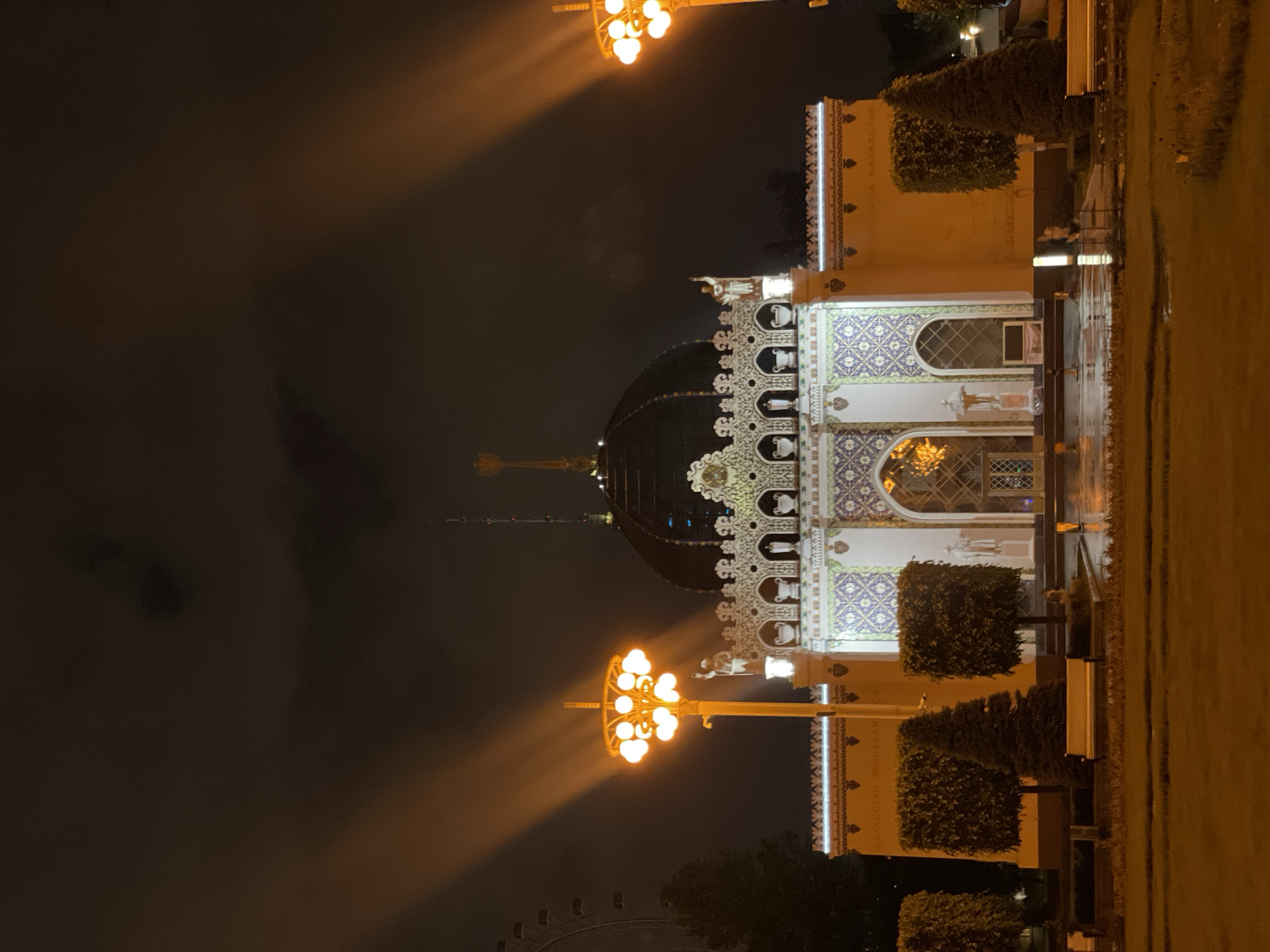 Captured at night in Moscow, Russia, the image focuses on the Kazakhstan Pavilion at VDNH. The pavilion is illuminated by street lamps, highlighting its architectural details, including a dome. The ground is wet, suggesting recent rain. Adjacent to the pavilion are manicured bushes and trees, typical of urban park landscaping.