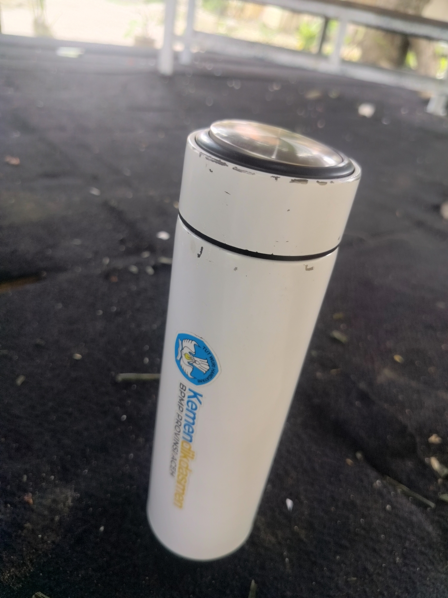 A white thermos with a silver lid rests on a dark, textured surface. A blue and white sticker with an emblem and text is affixed to the thermos. The background is blurred, showing hints of light-colored structures and foliage, suggesting an outdoor or semi-outdoor setting. The thermos appears to be positioned in the city of Purwodadi, Indonesia.