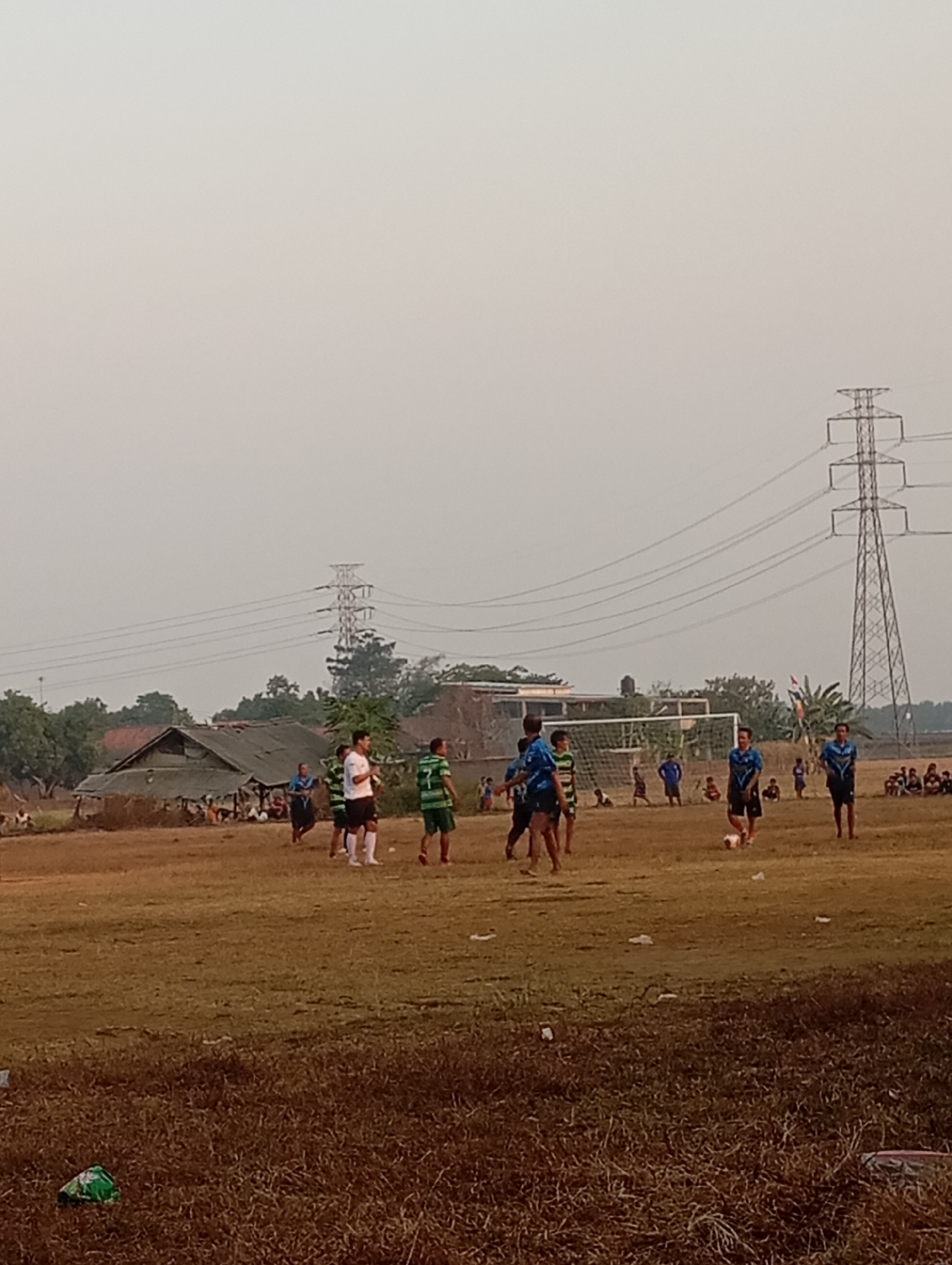 In Kecamatan Haurgeulis, Indonesia, a group of young men are engaged in a soccer match on a dry, dusty field. The game is being played in the late afternoon, with the sun casting long shadows. The field is surrounded by sparse vegetation and a few scattered buildings, including a dilapidated structure with a corrugated metal roof. In the background, a tall electricity pylon looms over the scene, its wires stretching across the hazy sky. Several spectators, likely locals, are scattered around the perimeter of the field, observing the game. The players are dressed in various colored jerseys and shorts, with some wearing cleats and others barefoot. The overall atmosphere appears casual and communal, indicative of a local sporting event in a rural Indonesian setting.