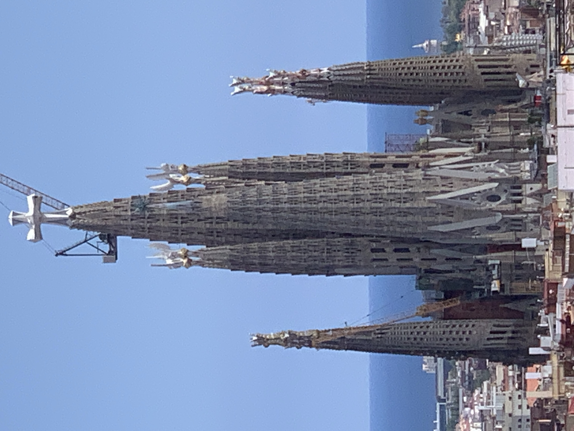 The image shows a daytime, exterior, medium shot of the Sagrada Familia basilica in Barcelona, Spain, with a clear blue sky and the Mediterranean Sea in the background. The basilica, a UNESCO World Heritage site, is a large, unfinished Roman Catholic minor basilica designed by Catalan architect Antoni Gaudí. The image focuses on several of its towering spires, which are intricately decorated with a variety of architectural details and sculptural elements. One of the spires is topped with a white cross, and a construction crane is visible near it, indicating ongoing construction or restoration. The lower parts of the image reveal a dense urban landscape of buildings with red-tiled roofs, characteristic of Barcelona's cityscape. The overall scene captures the scale and unique architectural style of the Sagrada Familia amidst its urban surroundings.