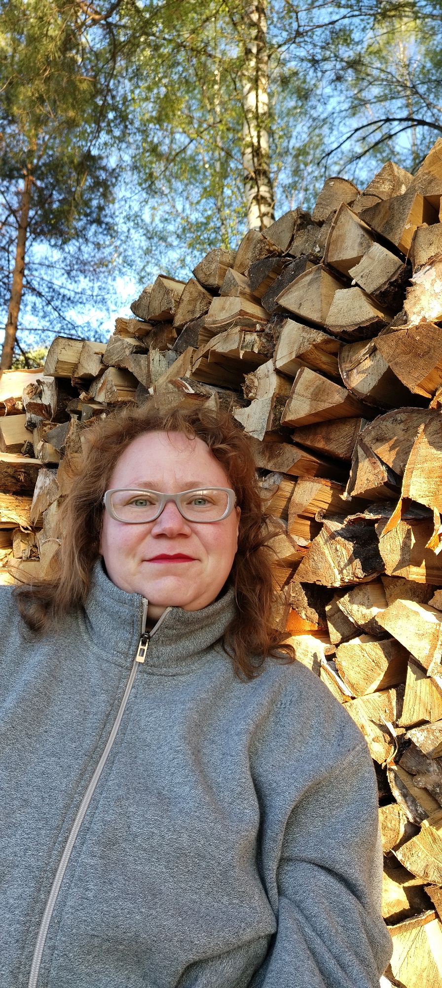 A woman with red hair and glasses is standing in front of a large pile of firewood. She is wearing a gray fleece jacket. The background shows trees and a blue sky. There is no information suggesting this image was taken in Зипуново, Russia.