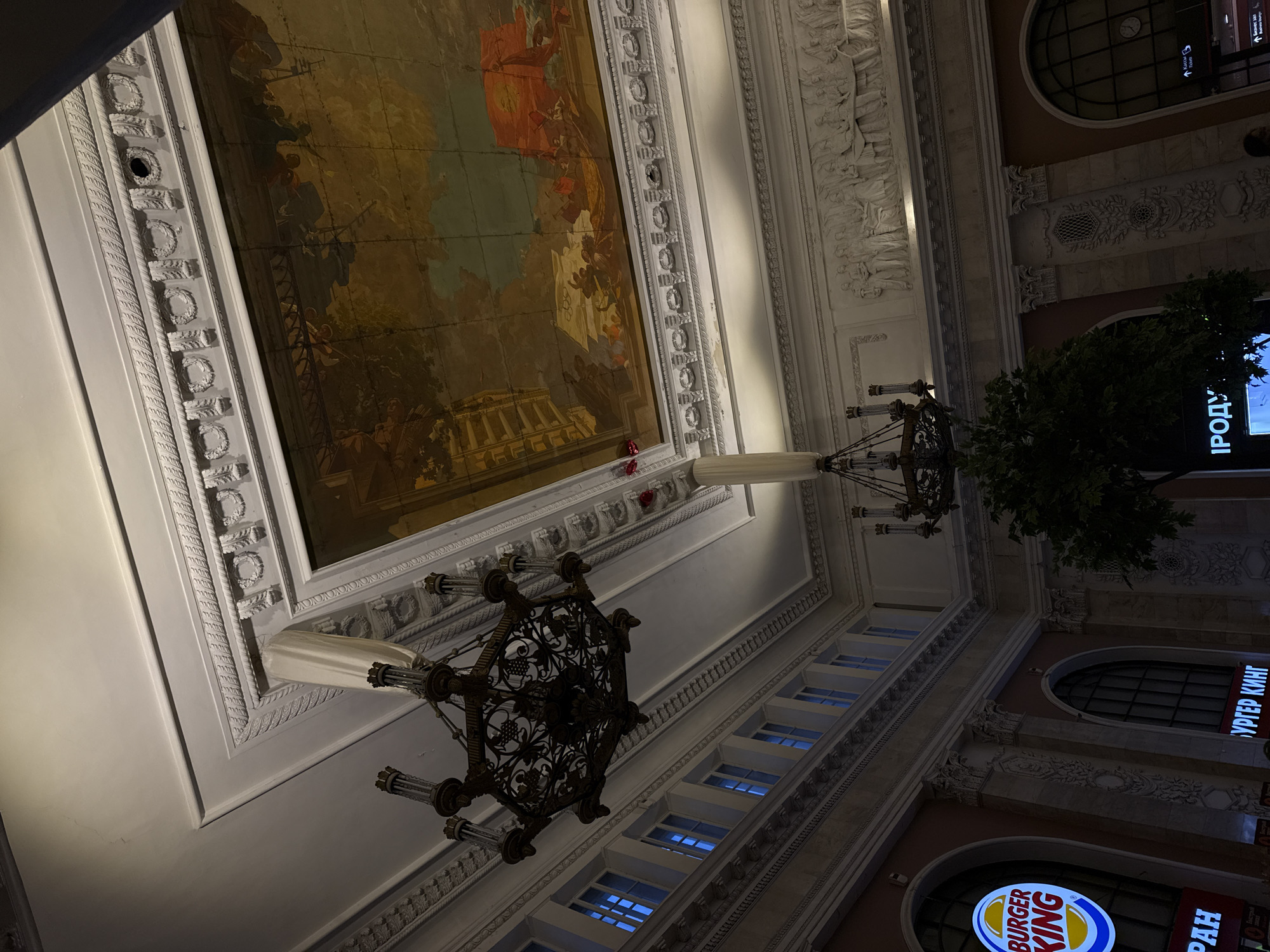 This media file presents an upward-angled view inside a grand, ornate building, likely in the evening or at night. The scene is dominated by a high ceiling or upper wall adorned with a large, faded fresco or mural depicting classical scenes, including figures and a temple-like structure. This mural is framed by elaborate white architectural moldings, cornices, and pilasters, intricately carved with laurel wreaths, geometric patterns, and classical relief sculptures featuring figures.
Two large, dark, ornate chandeliers, possibly made of wrought iron or bronze, hang prominently from the ceiling, featuring intricate, vine-like patterns and multiple light-holding arms.
Below these elaborate architectural details, the lower part of the scene reveals a juxtaposition of historical architecture with modern commercial elements. Rows of rectangular windows glow with a blue light, while arched windows are visible further down. Prominently displayed are illuminated signs, including the "BURGER KING" logo in English and Russian ("БУРГЕР КИНГ"). Other visible Russian text includes "ИРОДУ…" (a partial word), "Kacca" (Cashier), "Туалет" (Toilet), "Бизнес зал" (Business Lounge), and "Билетные кассы" (Ticket offices), indicating a public space like a train station or large commercial center. A small tree or large plant is also visible near one of the chandeliers and signs. No people are visible, and no specific activity is taking place within the frame. The artificial lighting suggests it's nighttime.