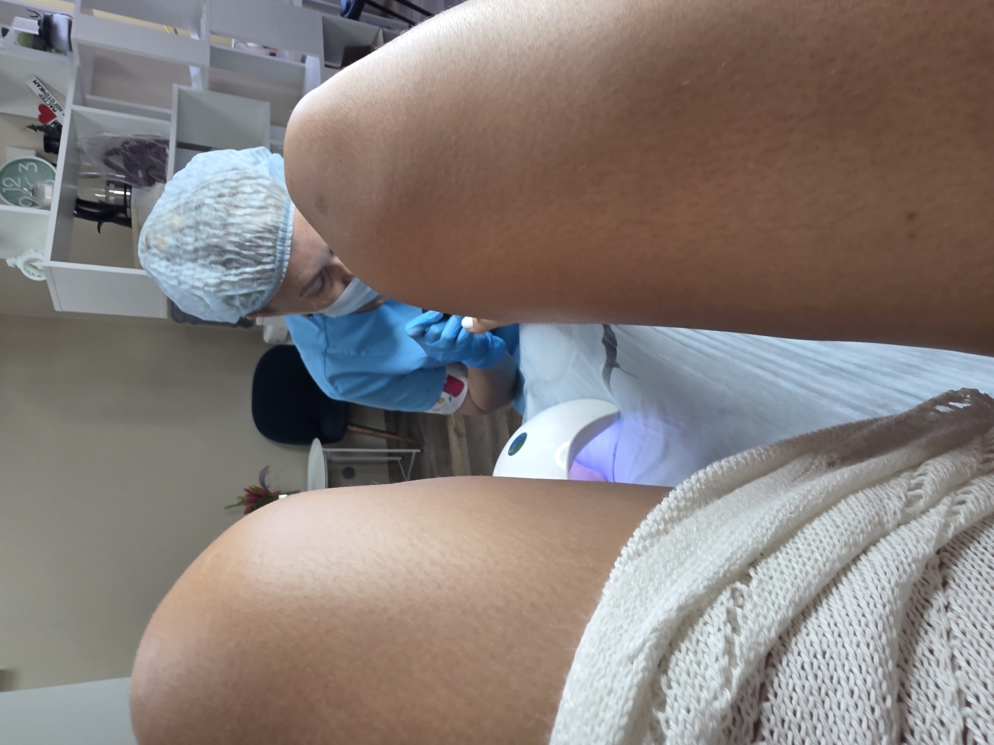 In a nail salon, a person in a light blue uniform and hairnet is applying nail polish to another person's foot, which is resting on a white towel near a UV nail lamp. The person receiving the treatment is wearing a cream-colored knitted garment. The background shows a shelf with decorative items, including a clock and a framed picture. The location is identified as Sochi, Russia.