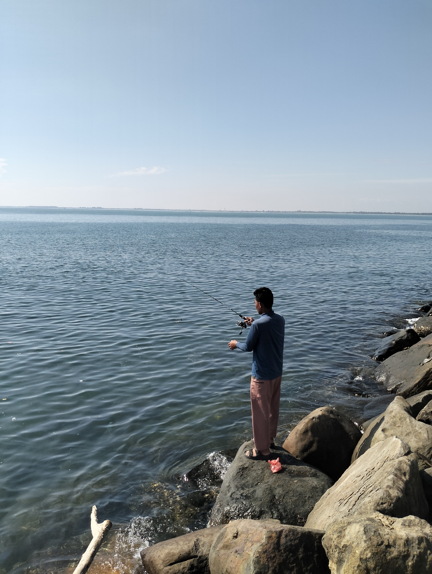 A man is fishing in Lhokseumawe, Indonesia. He stands on a pile of large, gray rocks that line the edge of the water. He wears a blue, long-sleeved shirt, salmon-colored pants, and sandals. He holds a fishing rod, casting a line out into the calm water. The water is a deep blue, and the sky above is clear and pale blue. In the distance, the shoreline is visible. A piece of driftwood lies on the rocks in the lower left corner of the image. There is a red plastic bag on the rocks near the man's feet.