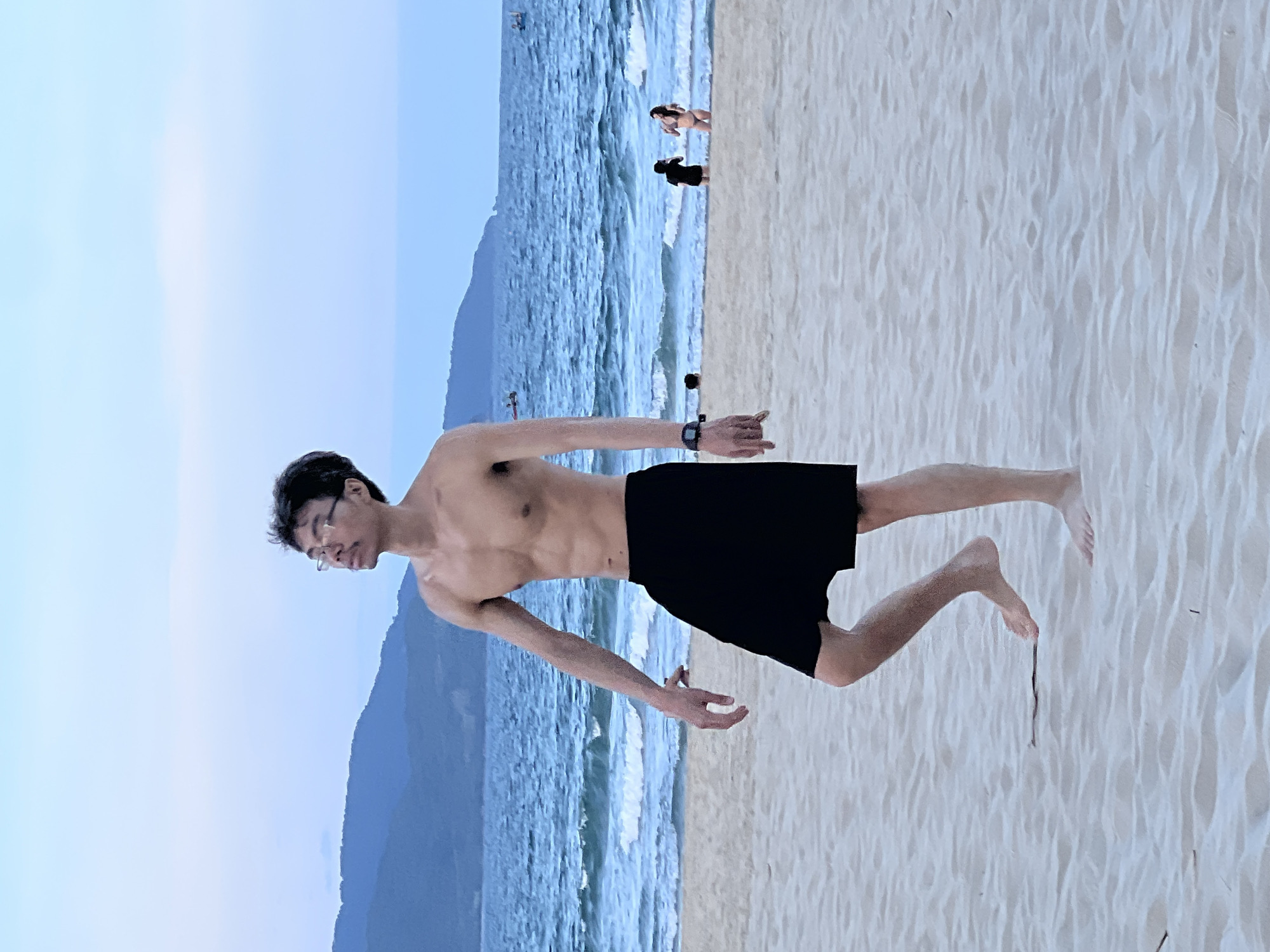 A young man with glasses walks on a sandy beach in Đà Nẵng, Vietnam. He is shirtless, wearing black shorts, and looking upwards with his mouth slightly open. His arms are extended downwards and outwards. The ocean is behind him, with waves rolling onto the shore. In the distance, mountains are visible against a pale sky. Other people are scattered on the beach and in the water, some wading and others standing. The sand is light in color and shows ripples from the wind. A dark object, possibly a piece of debris, lies on the sand in front of the man's left foot.