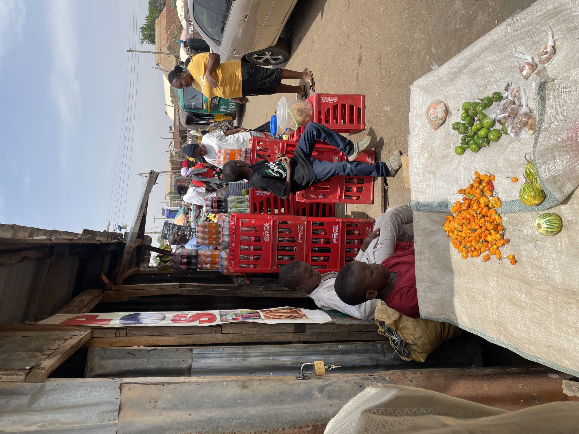 The image captures a scene in Jos, Nigeria, depicting a bustling street market. In the foreground, two boys are resting on a platform or wooden surface next to a market stall with a bag underneath. A table covered with a white cloth displays an assortment of vegetables and spices including yellow peppers, green eggplants, wrapped spices and garlic. Above them, a boy is relaxing on a pile of red Coca-Cola crates, with a woman in a yellow shirt standing nearby. Visible behind the crates are various soft drinks and other goods, suggesting this area is a vendor's stand. In the background, more people are visible among other stalls and a car. The setting appears to be an open-air market, with makeshift structures providing shade and shelter for the vendors and their wares. The environment suggests a lively commercial activity.