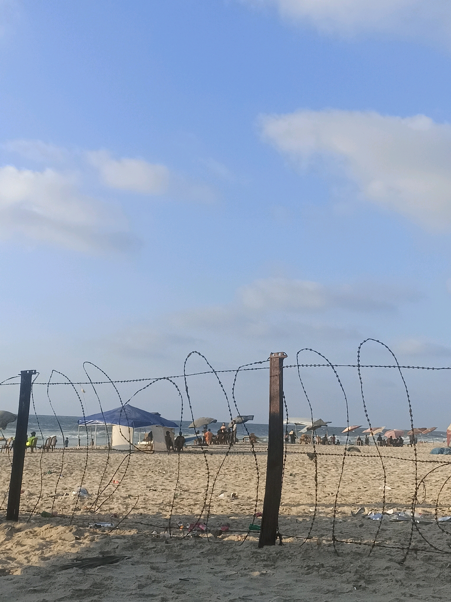 The image depicts a view of a beach in Gaza, Palestinian Territories, behind a barrier of barbed wire. The foreground is dominated by the sand and the rusty barbed wire fence, with some debris scattered on the ground. In the background, the sea stretches to the horizon under a bright blue sky with scattered white clouds. Several people are visible on the beach, some under beach umbrellas and tents, suggesting a public gathering or recreational activity. The presence of the barbed wire suggests a restricted or monitored area.