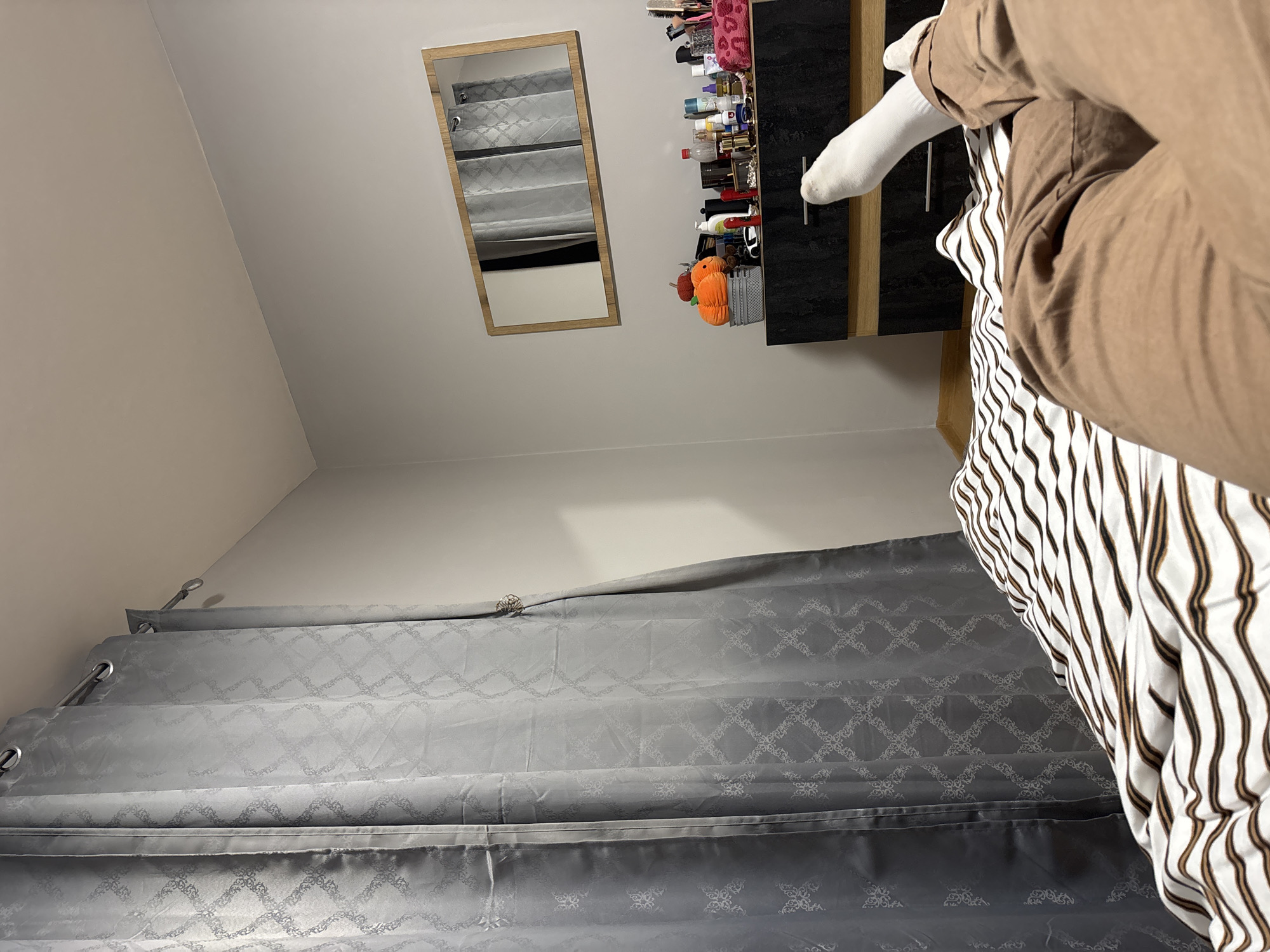 The image shows an interior scene with a gray curtain covering the lower portion of the frame. Above the curtain, the wall is a light color. On the right side, there is a person wearing a light brown outfit, a white sock, and a striped shirt. On the upper part of the image, there's a wooden-framed mirror and a cabinet with various items on its shelves. The location of Burgas, Bulgaria is not evident in the scene as no specific elements from the city are visible.