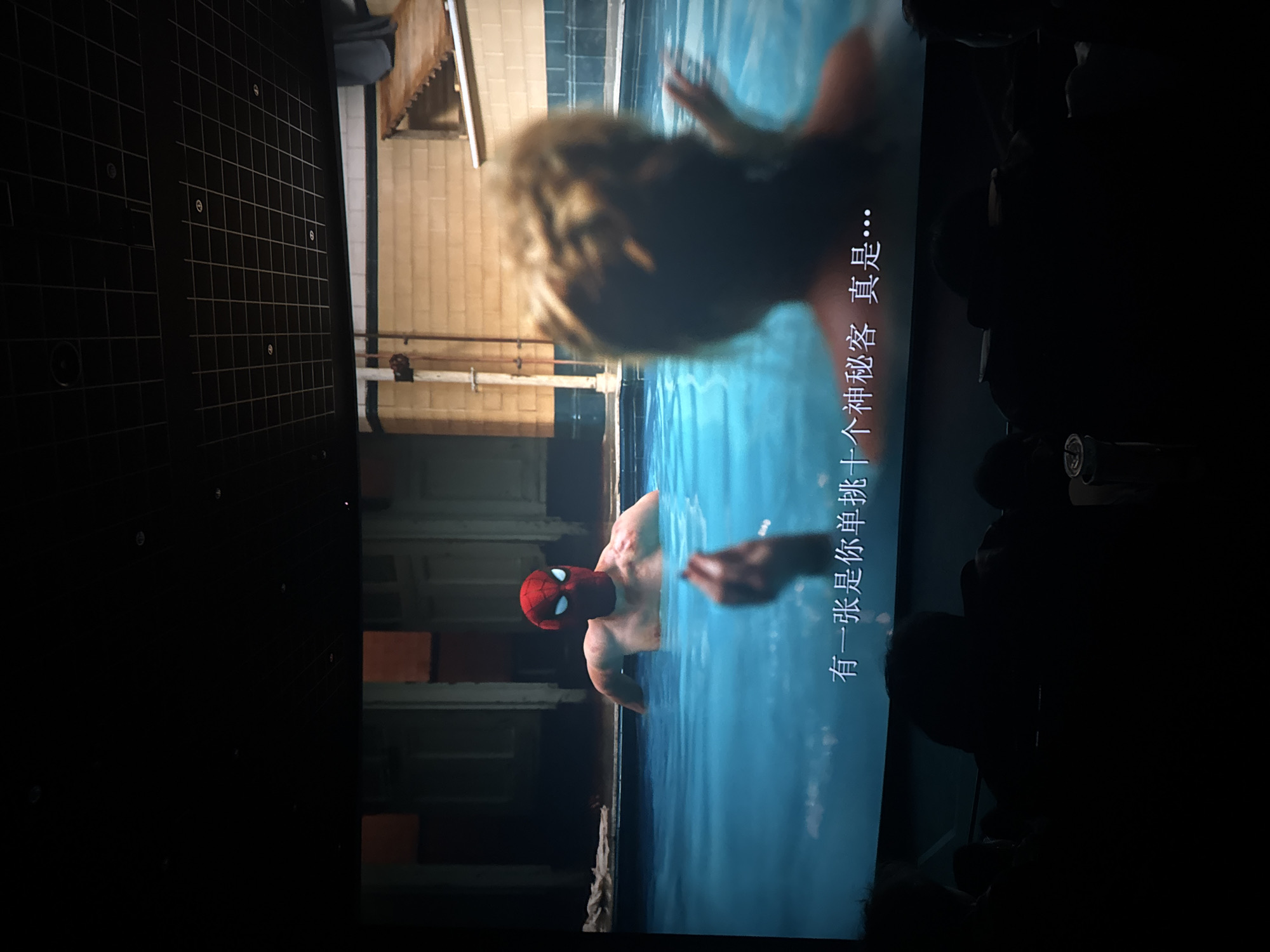 The image shows a movie scene displayed on a screen. In the foreground, a person wearing a Spider-Man mask is submerged in blue water, with only their head and shoulders visible. Their body is muscular, and they appear to be looking towards the right side of the frame. In the background, another person, mostly out of focus, is also in the water, with their back to the camera. The setting appears to be an indoor swimming pool or a similar body of water, with tiled walls and pipes visible. There is text in Chinese characters on the right side of the screen, which reads "有一张是单挑十个神秘客真是…". This translates to "There is one who challenges ten mysterious guests, truly...". The overall lighting is dim, suggesting a cinematic presentation or a dimly lit room.
