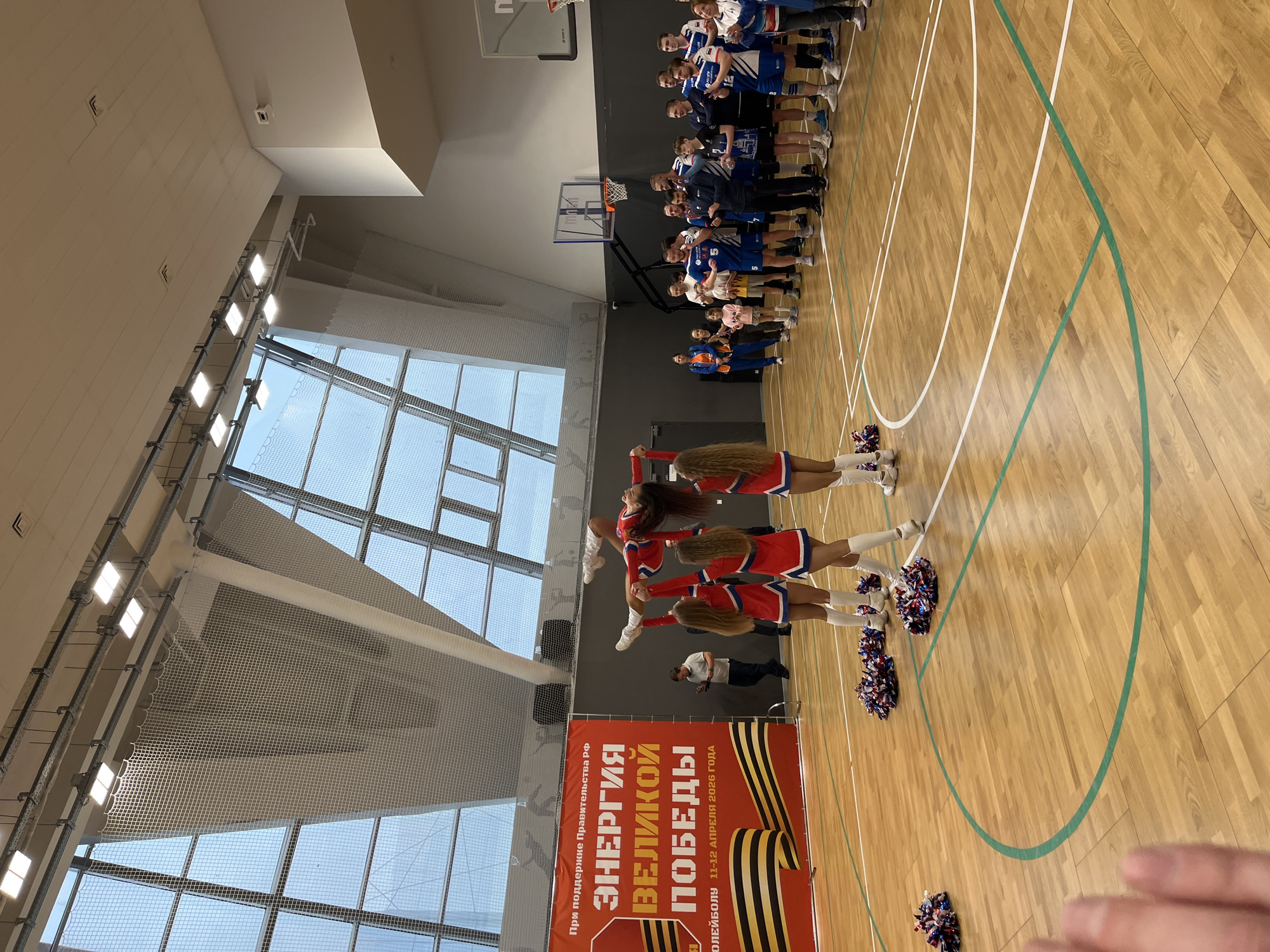 A gymnasium is the setting for a photo taken in Moscow, Russia. Visible are cheerleaders, pom-poms, and a basketball team on the court. A promotional banner is present, and natural light is available via windows.